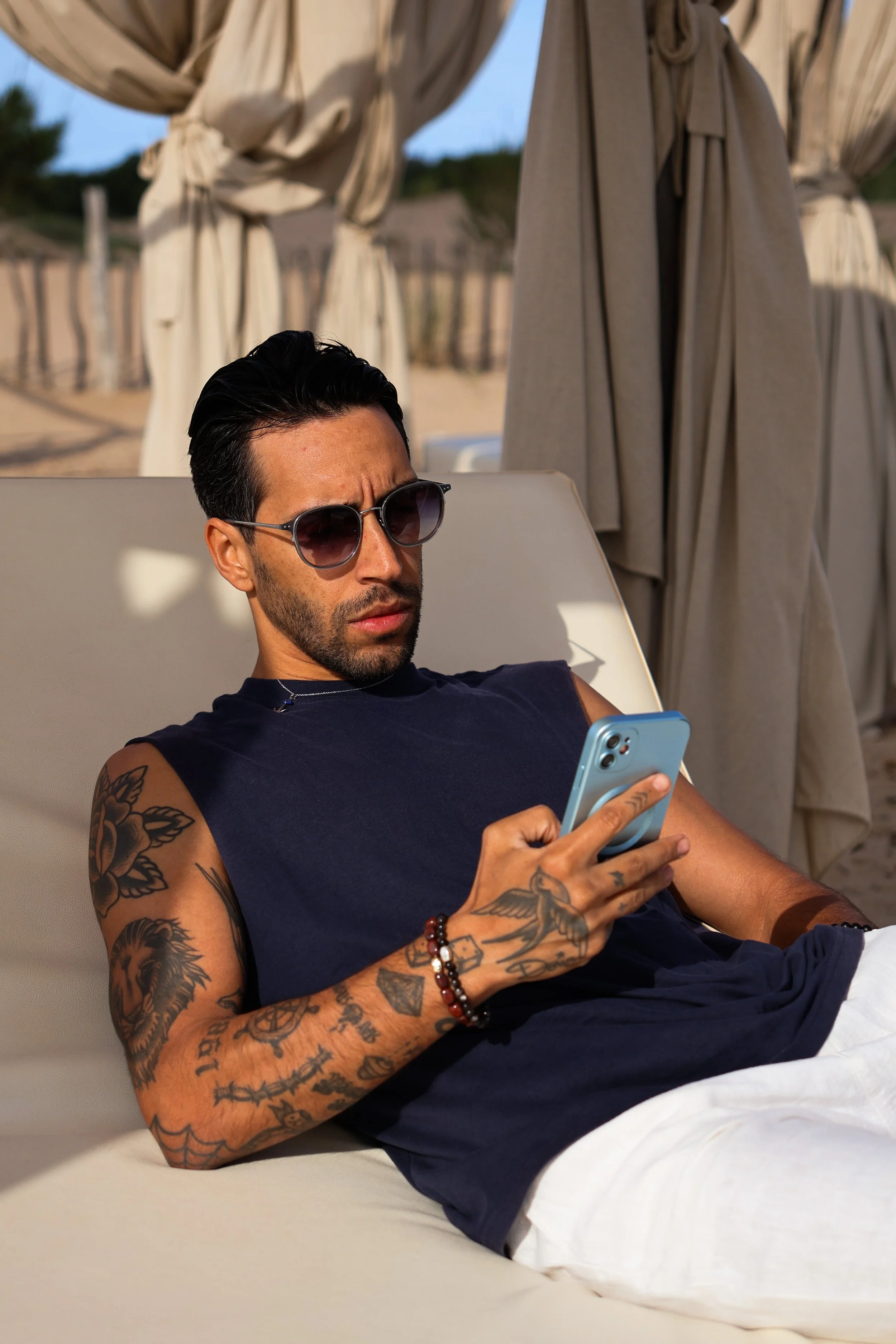 A man with dark hair, sunglasses, and tattoos on his arms relaxing on a lounge chair outdoors, looking at his smartphone.