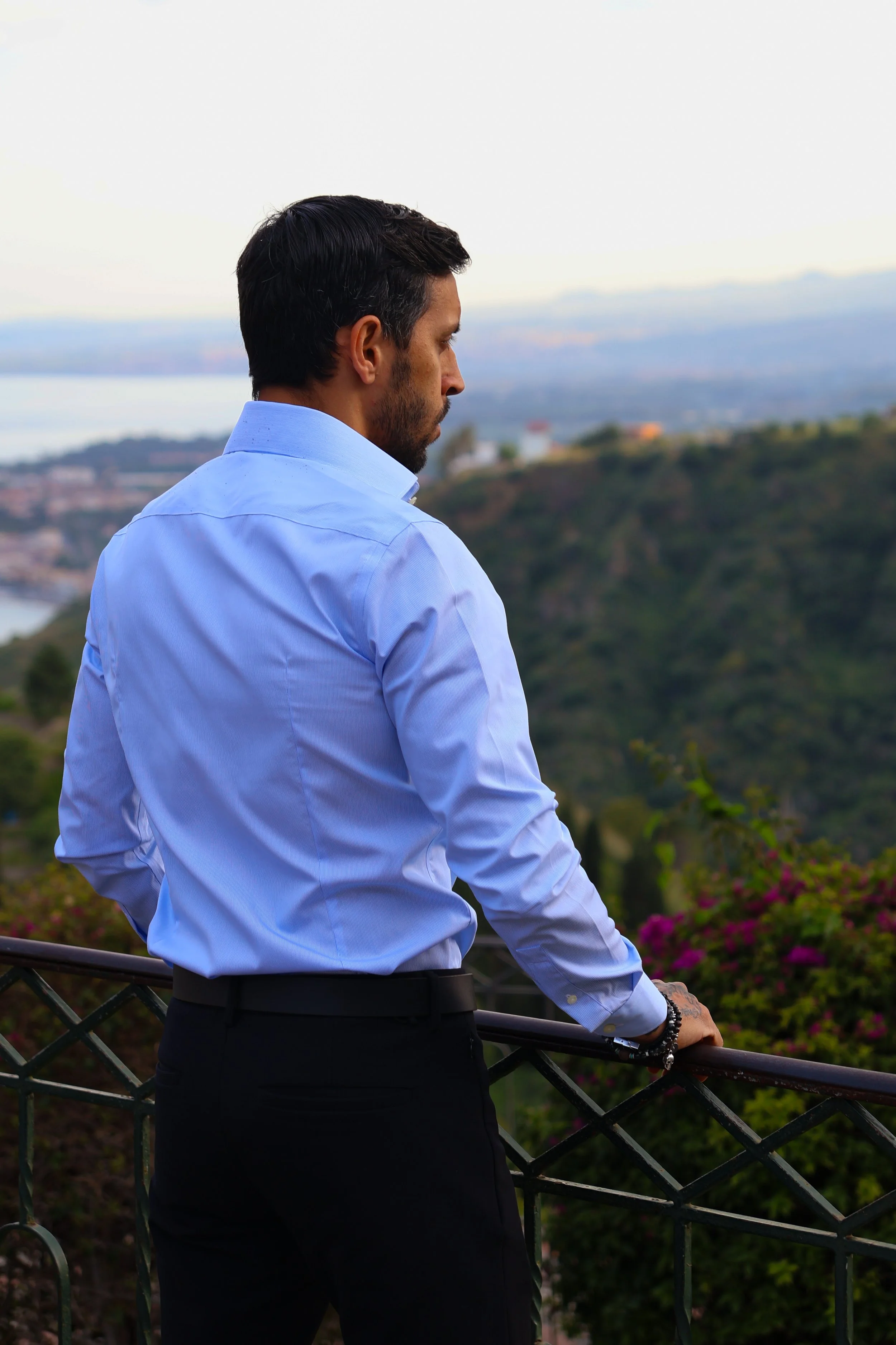 A man in a white dress shirt and black pants standing by a railing, looking out at a scenic landscape with hills, water, and distant buildings during sunset.