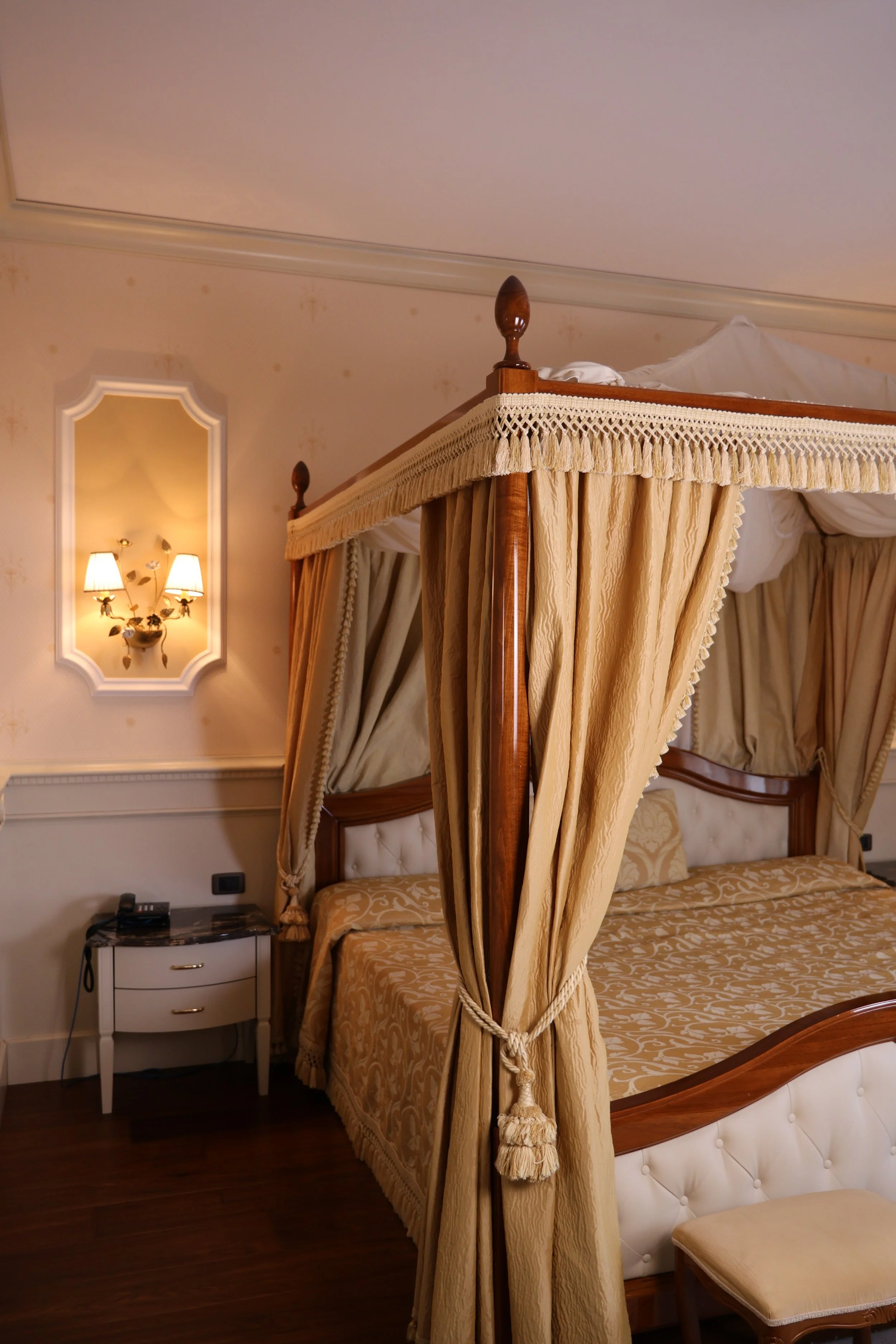 A luxurious four-poster bed with golden curtains and a tufted headboard, next to a small nightstand with a phone and lamps on the wall.