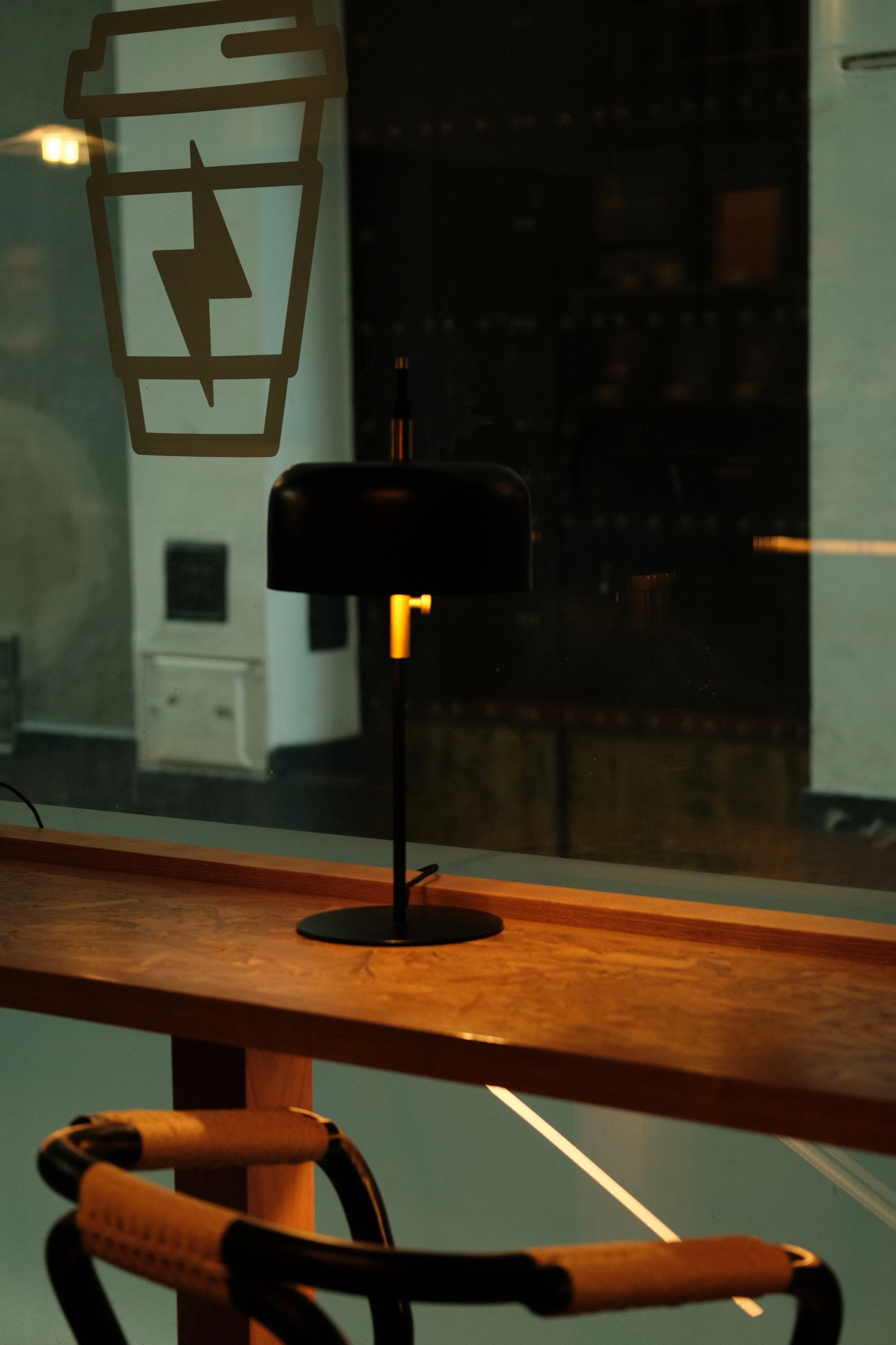 A black table lamp with a curved shade on a wooden table near a large window with a coffee cup symbol sticker. The background shows a dark interior space.