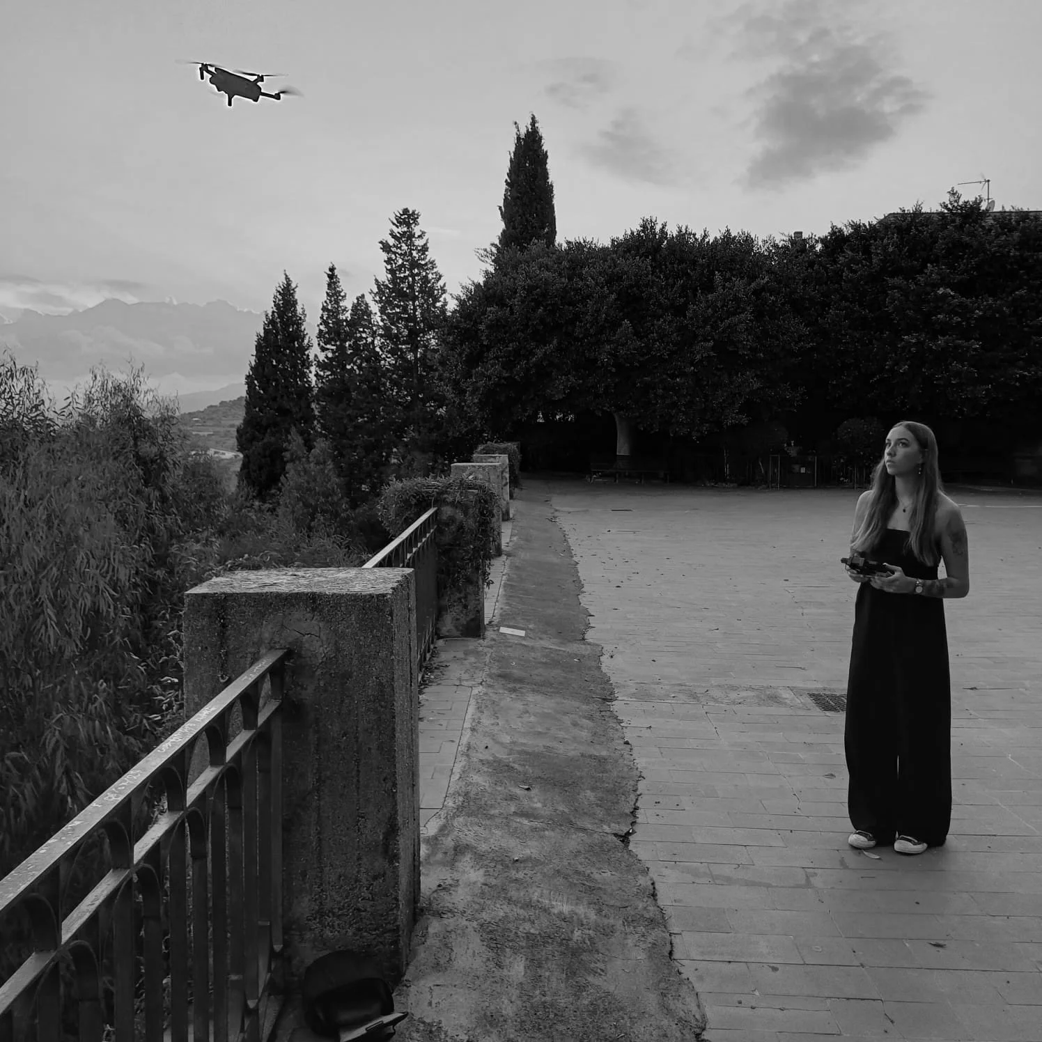 A woman in a black dress stands on a paved area holding a remote control, looking up at a flying drone in the sky. The scene is outdoors with trees and a mountain range in the background, and the photo is in black and white.