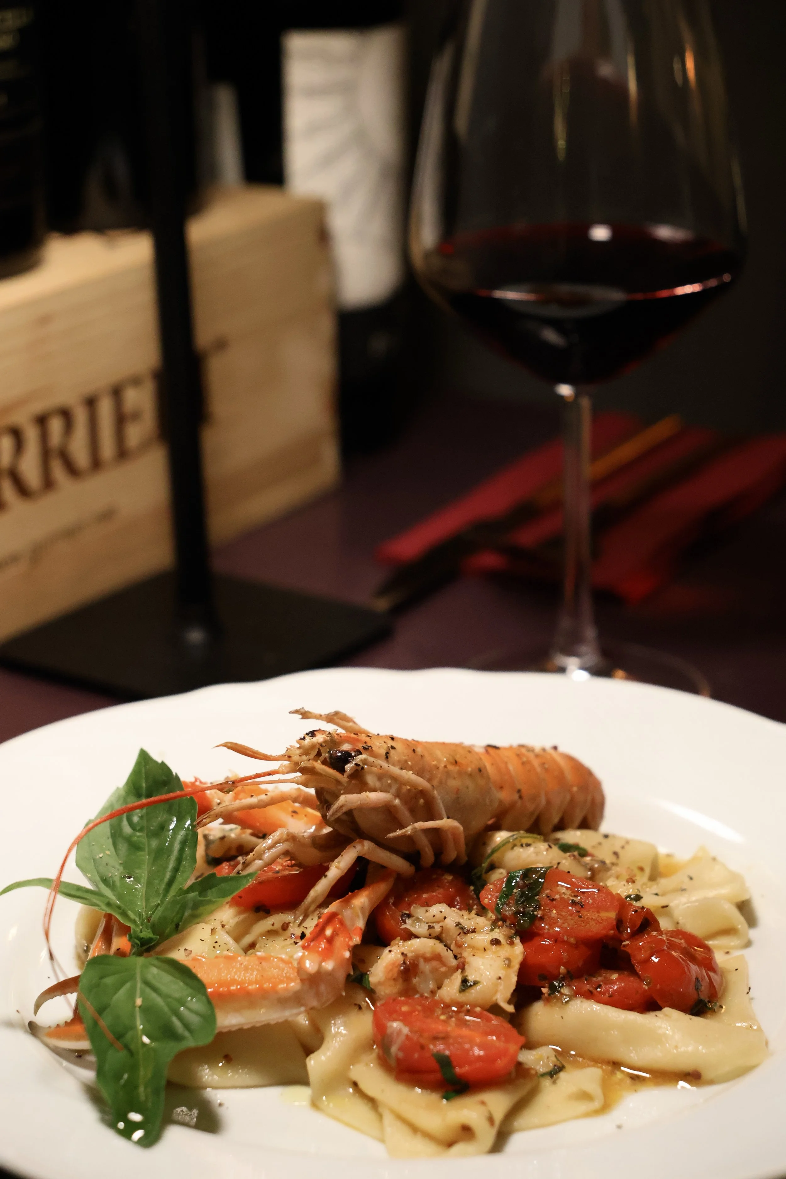 Plate of pasta with lobster, cherry tomatoes, and fresh basil, served with a glass of red wine in the background.