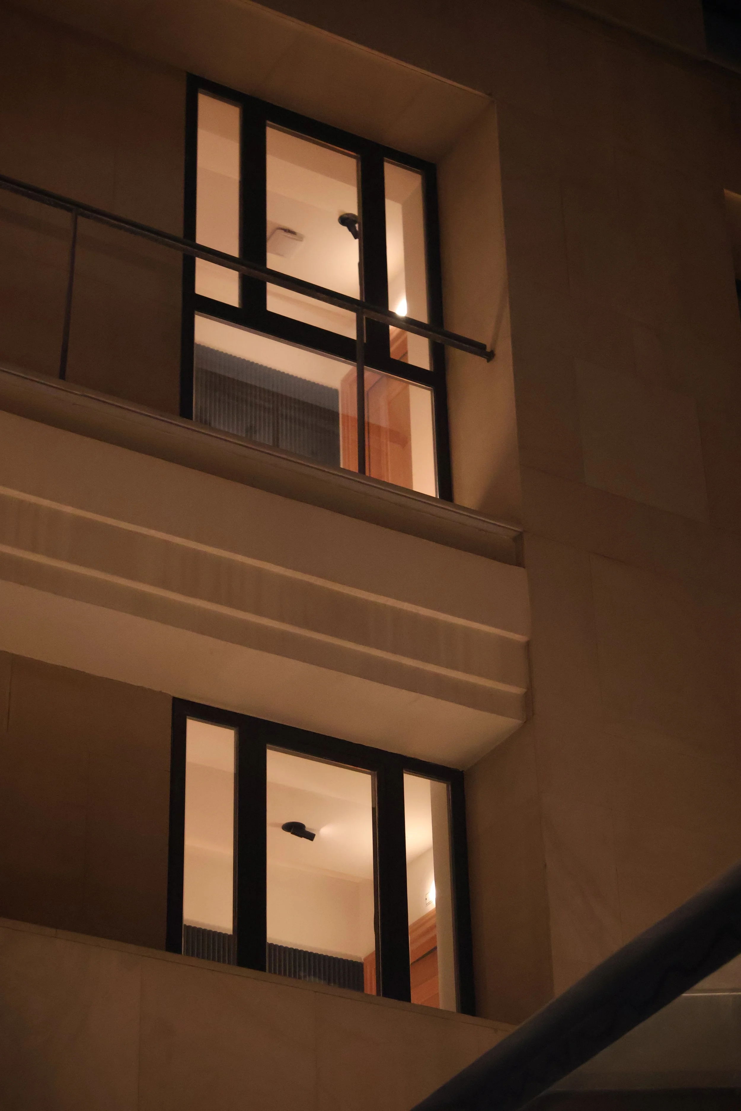 Nighttime view of a modern building with tall glass windows, showing illuminated interior rooms with ceiling lights, seen from an angle including part of a staircase railing.