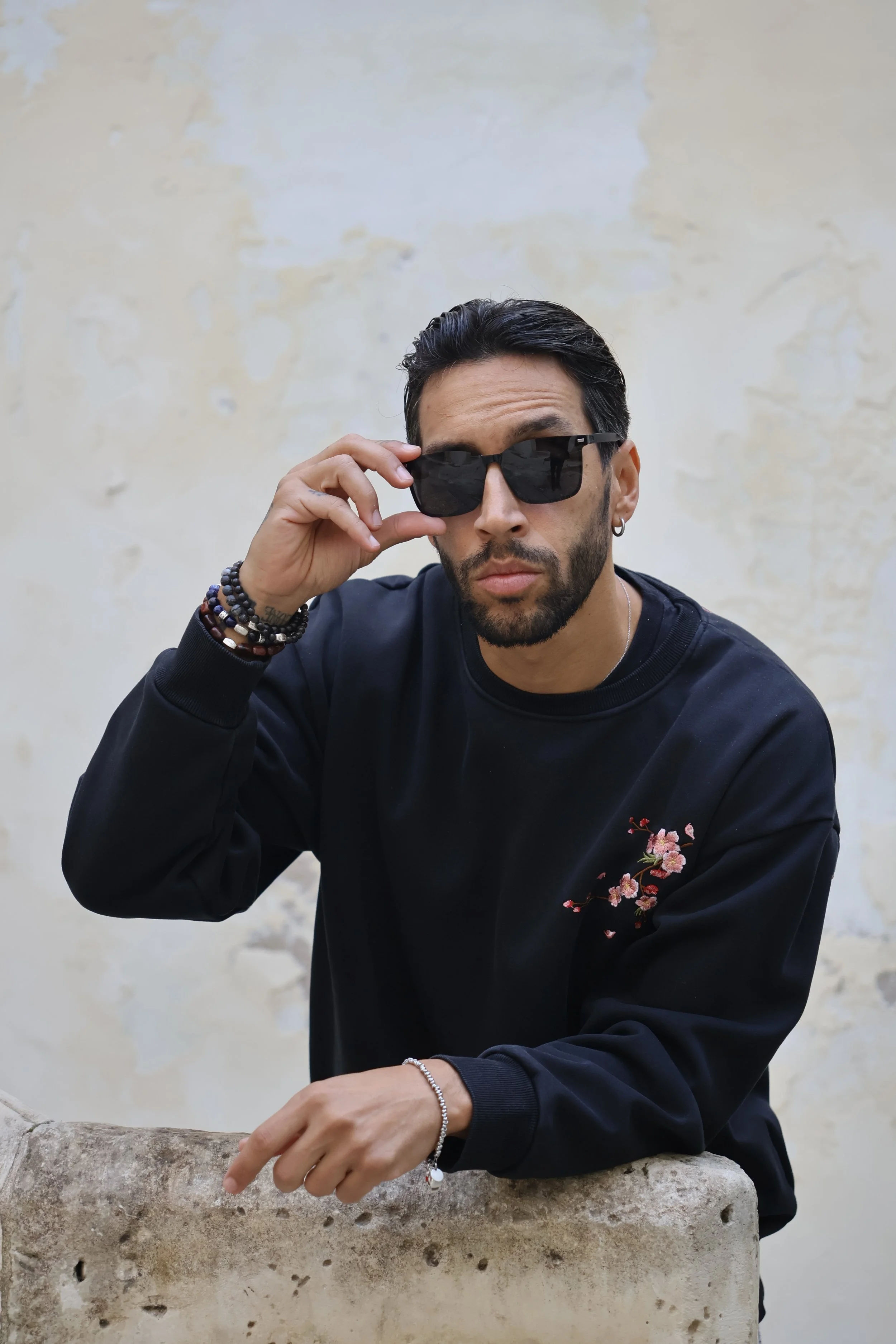 A man wearing sunglasses and a black sweatshirt with floral embroidery, leaning on a stone ledge against a weathered light-colored wall.