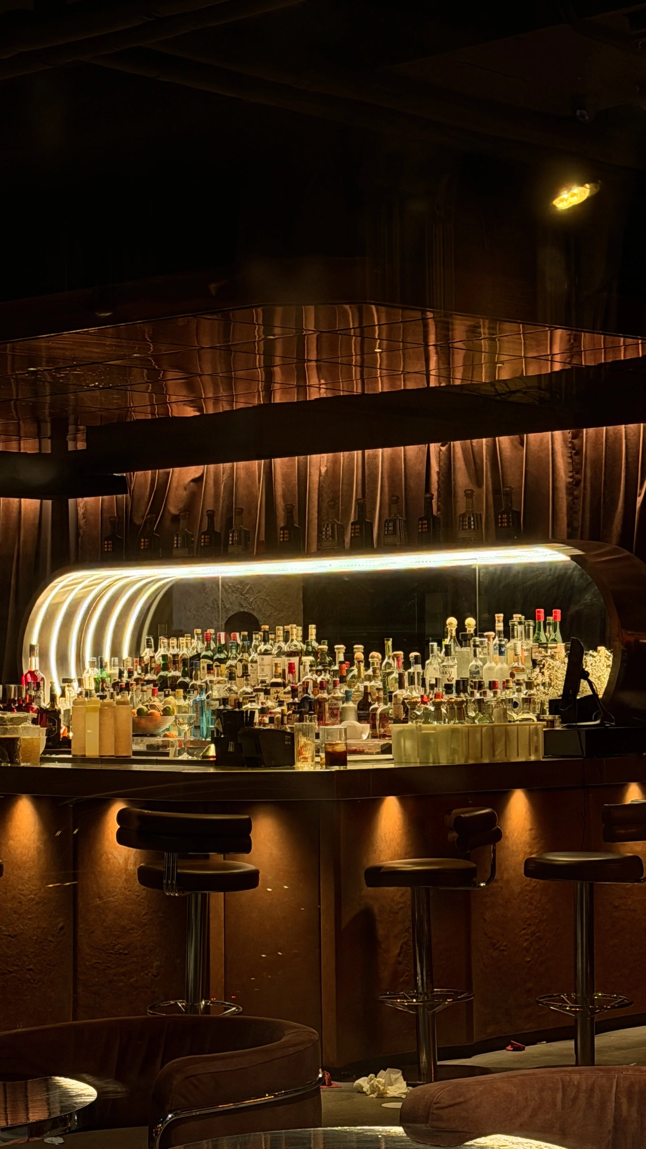 A dimly lit bar with a curved counter filled with various liquor bottles and glasses, illuminated by warm lights.