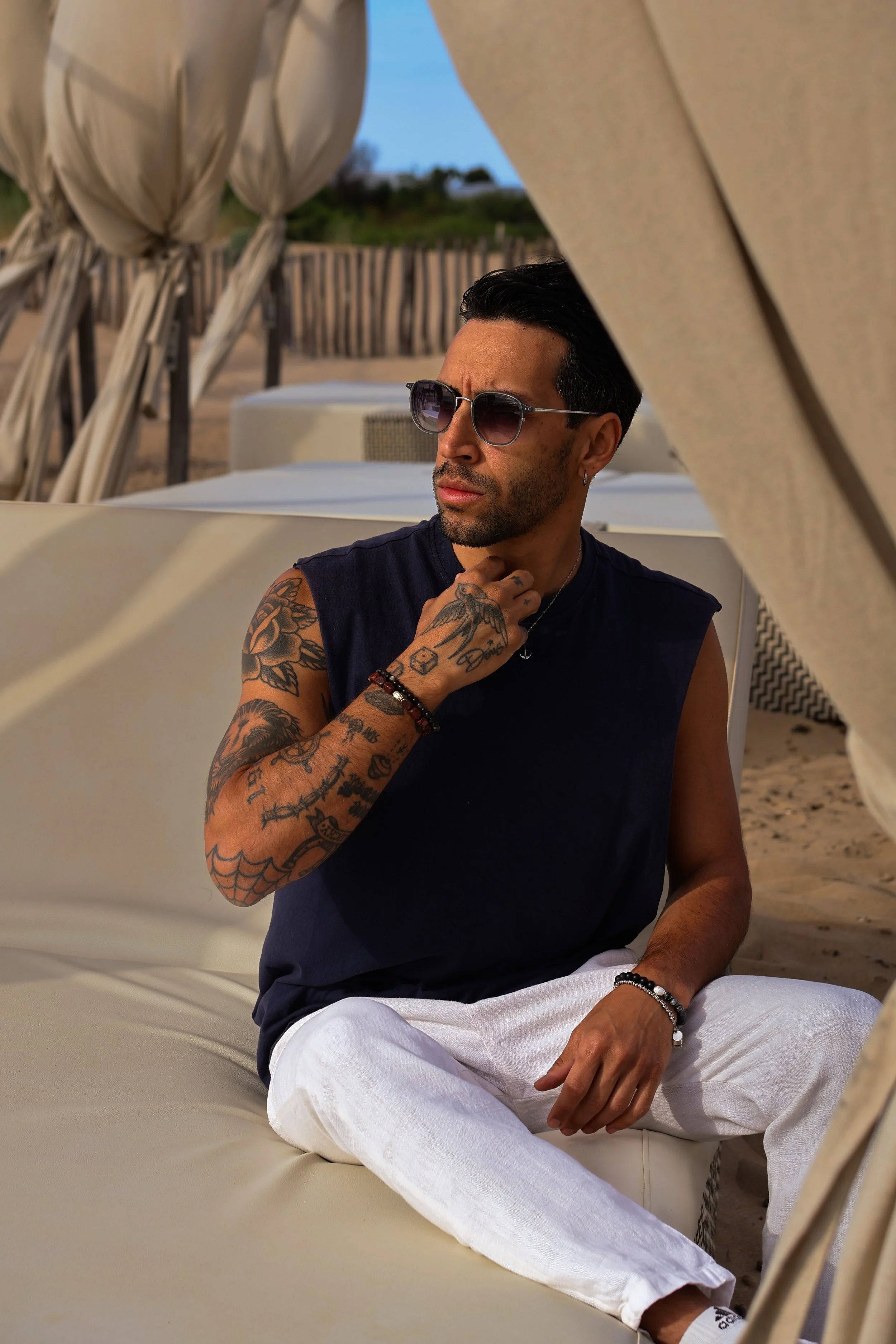 A man with tattoos on his arms and chest, wearing sunglasses, a black sleeveless shirt, white pants, and jewelry, sitting outdoors in a relaxed pose on a white sofa at a beach resort during the evening.