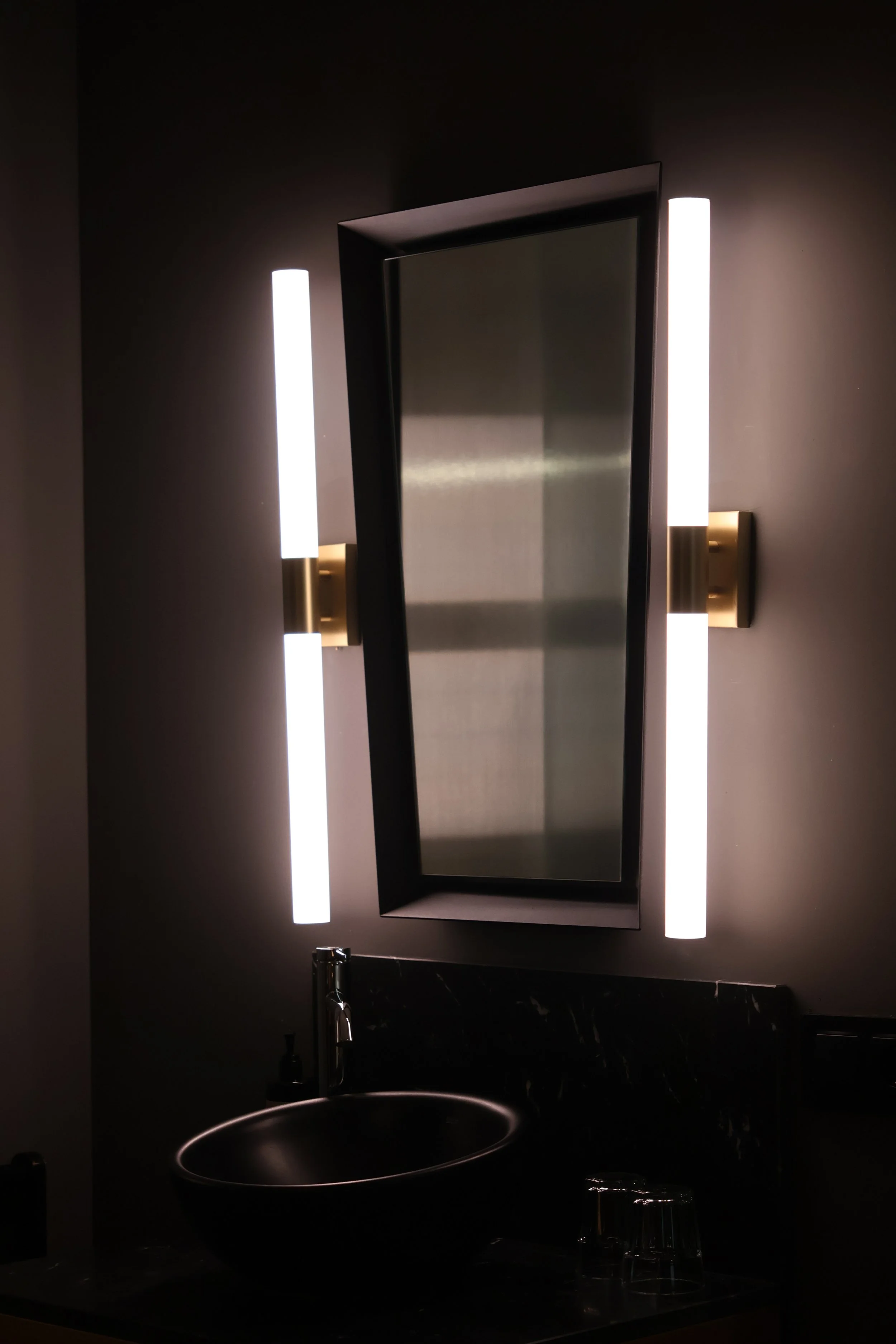Modern bathroom with a rectangular mirror, vertical LED lights on each side, a black vessel sink, and a dark countertop.