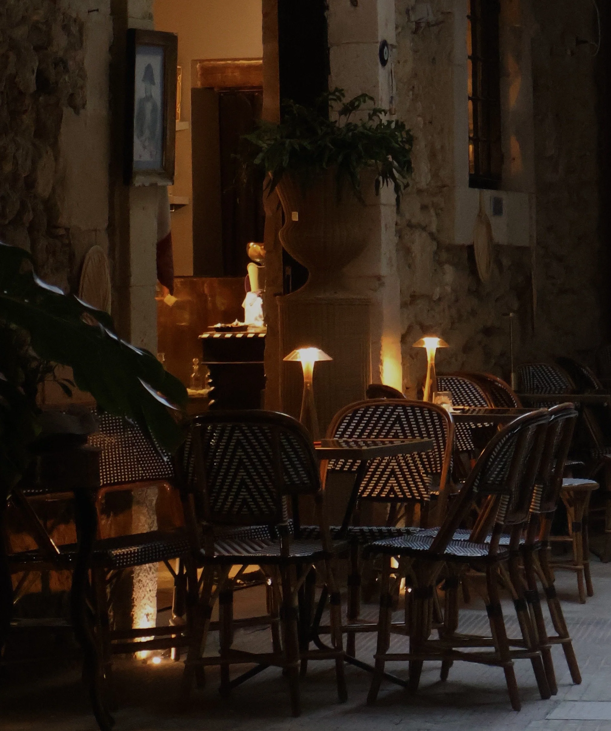 A cozy dining area with wicker chairs around a table, illuminated by small table lamps, with a large potted plant and stone walls in the background.