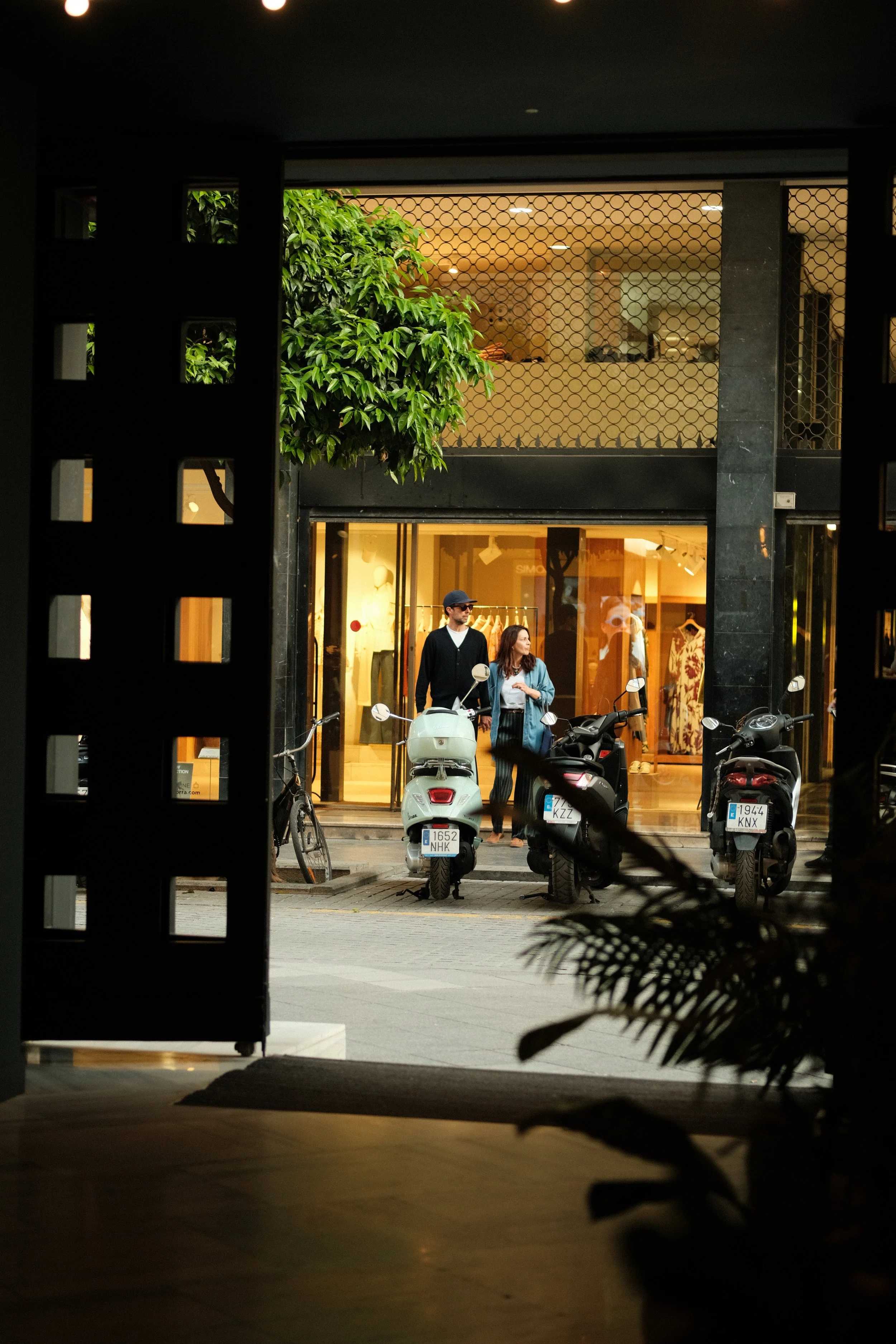 People walking outside a storefront at night, parking scooters and a bicycle on the sidewalk, with indoor lighting and mannequins seen through the glass.