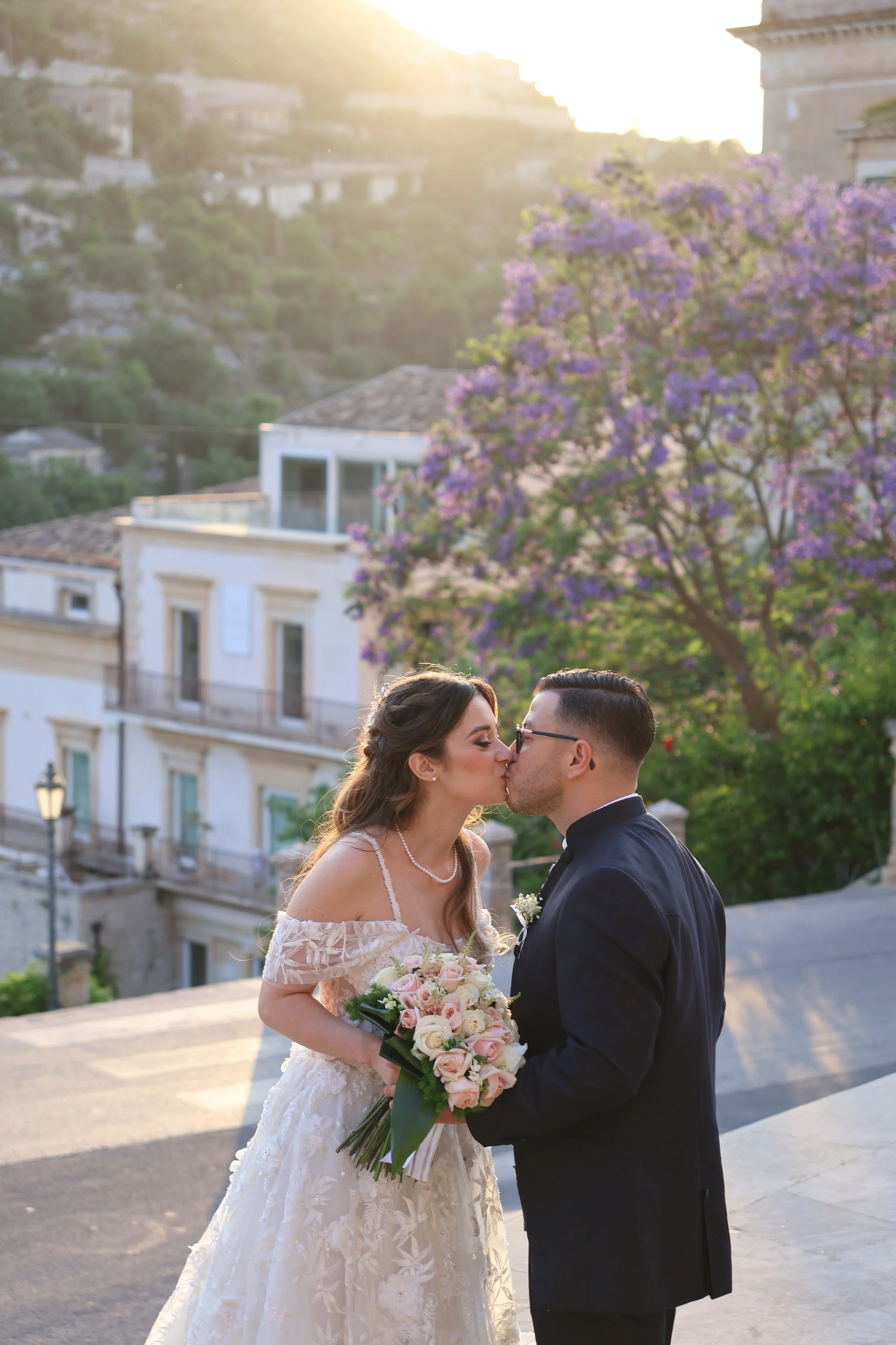 A newlywed couple sharing a kiss outdoors during sunset, with a woman in a wedding dress holding a bouquet of pink roses and a man in a black suit