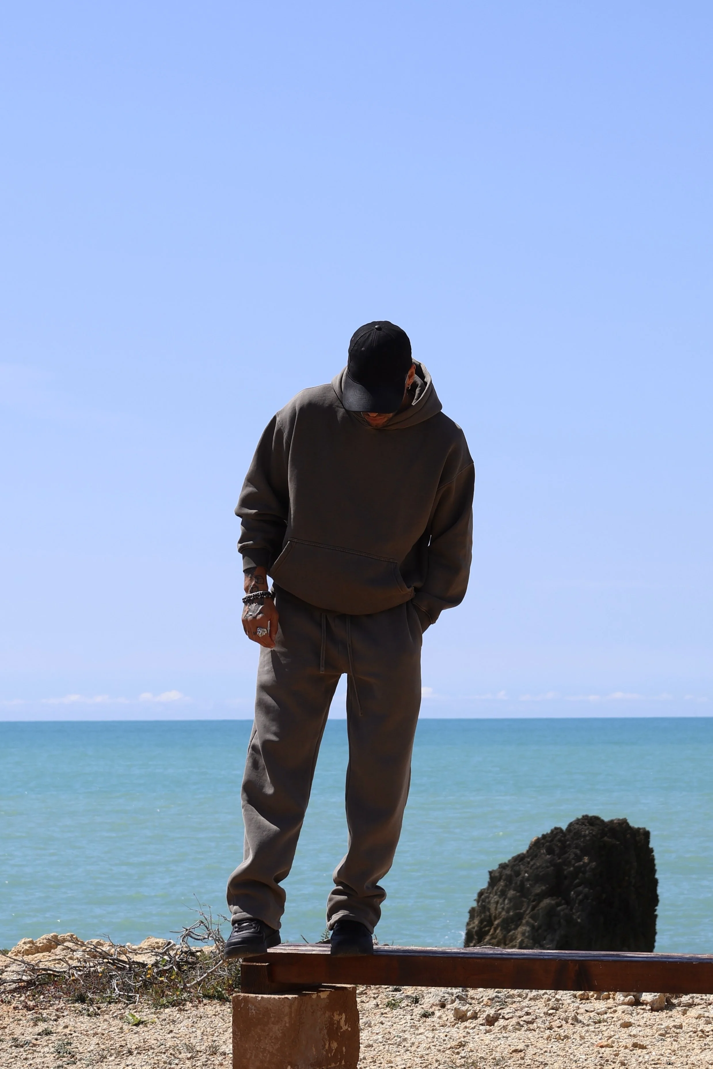 Editorial foto for men's fashion. Man wearing a black cap and gray hoodie standing on a wooden beam at the beach, with the ocean and blue sky in the background.