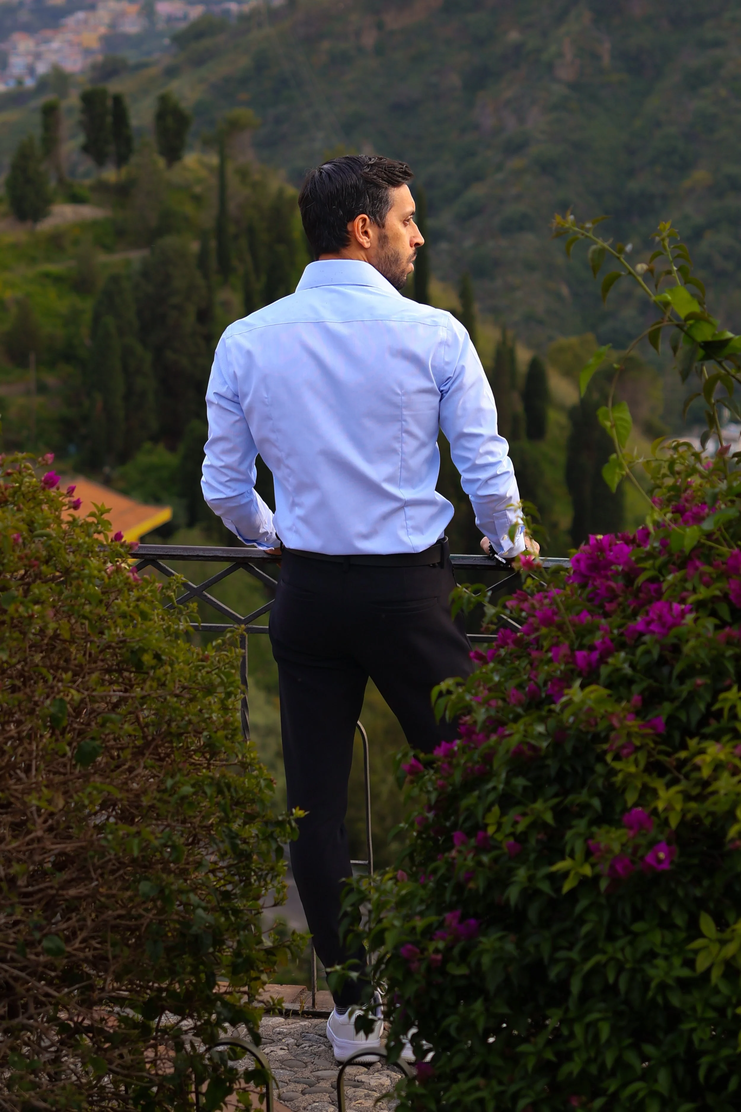 A man in a light blue shirt and black pants standing on a balcony, looking at a lush green landscape with trees and hills in the background, surrounded by flowering bushes.