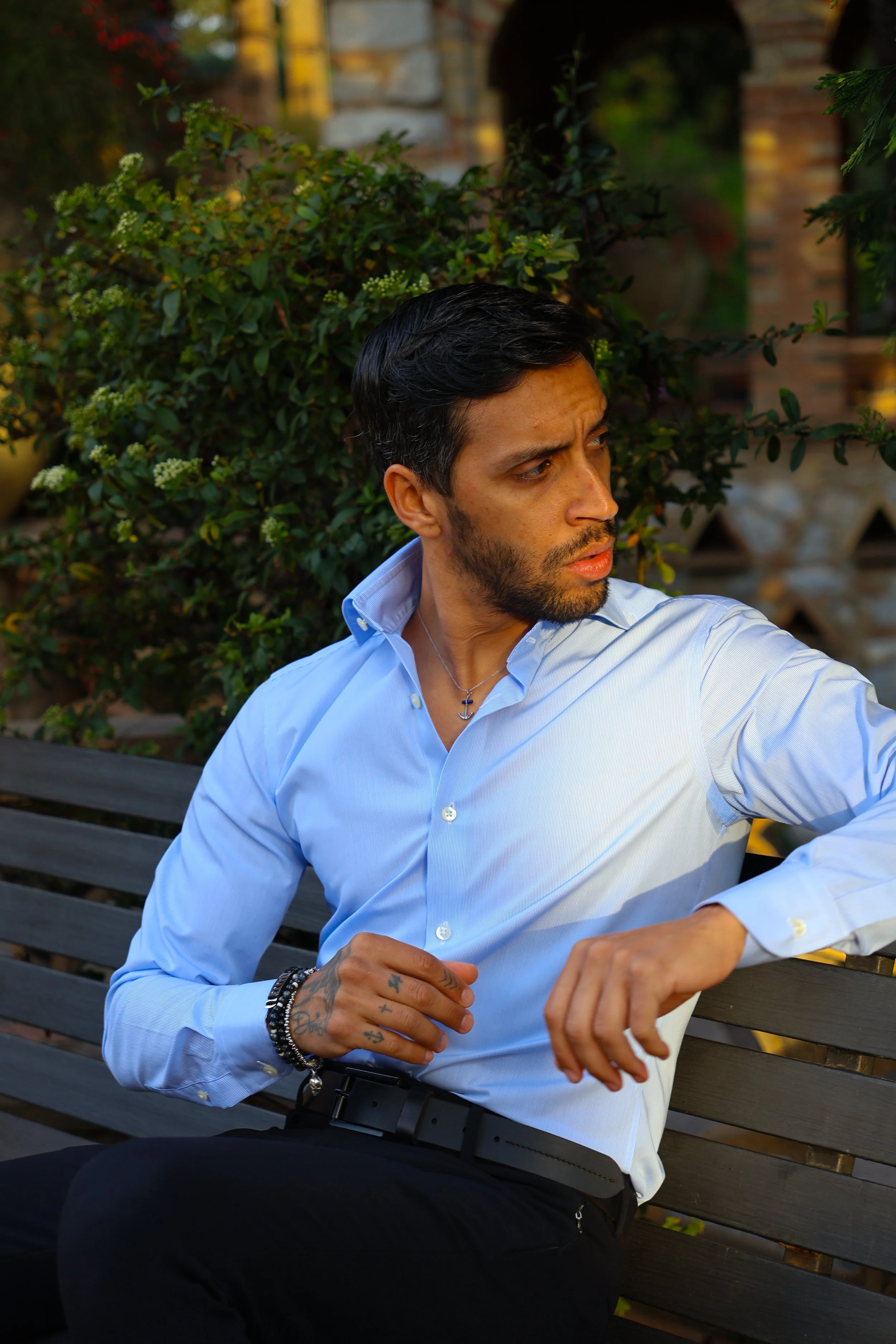 A man with dark hair and a beard wearing a light blue dress shirt and black pants sitting on a park bench, looking to his left, with greenery and a brick wall in the background.