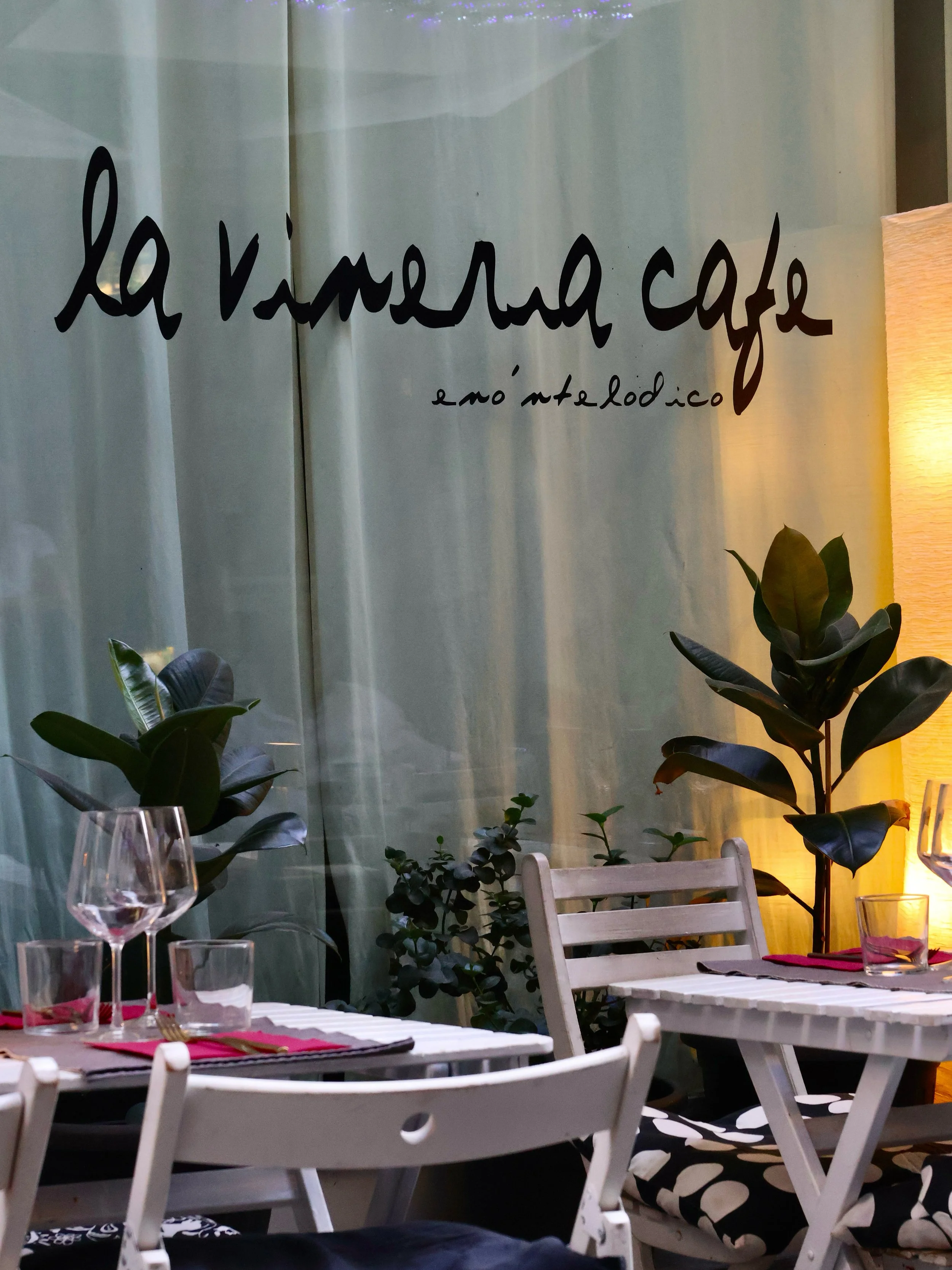 Exterior of La Vineria Cafe with tables, chairs, glassware, and potted plants, seen through a glass window with the cafe's name written onscreen.