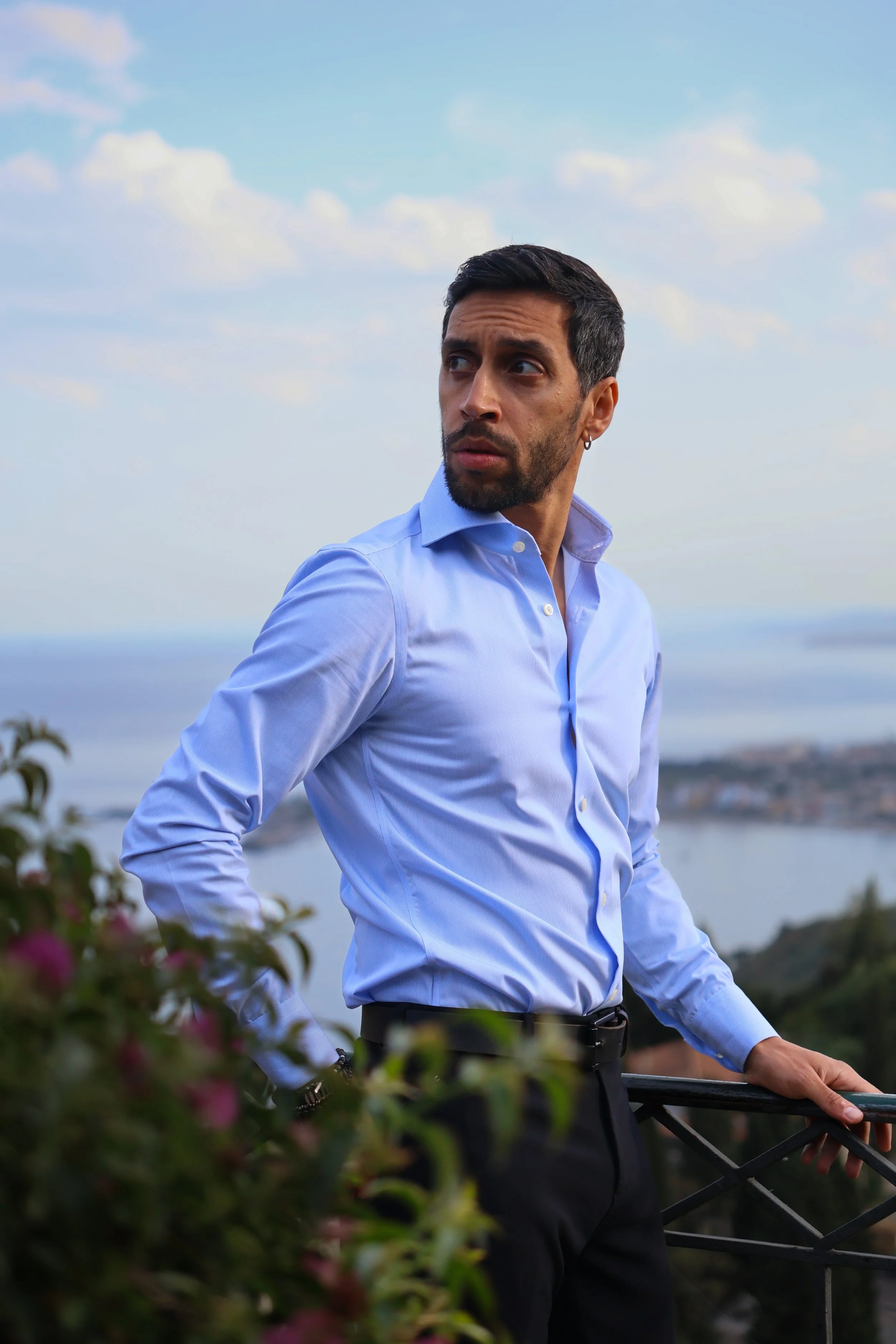 A man with dark hair, beard, and a small earring in his left ear, wearing a light blue dress shirt, stands outdoors on a balcony overlooking a landscape with water and distant shoreline, against a partly cloudy sky.