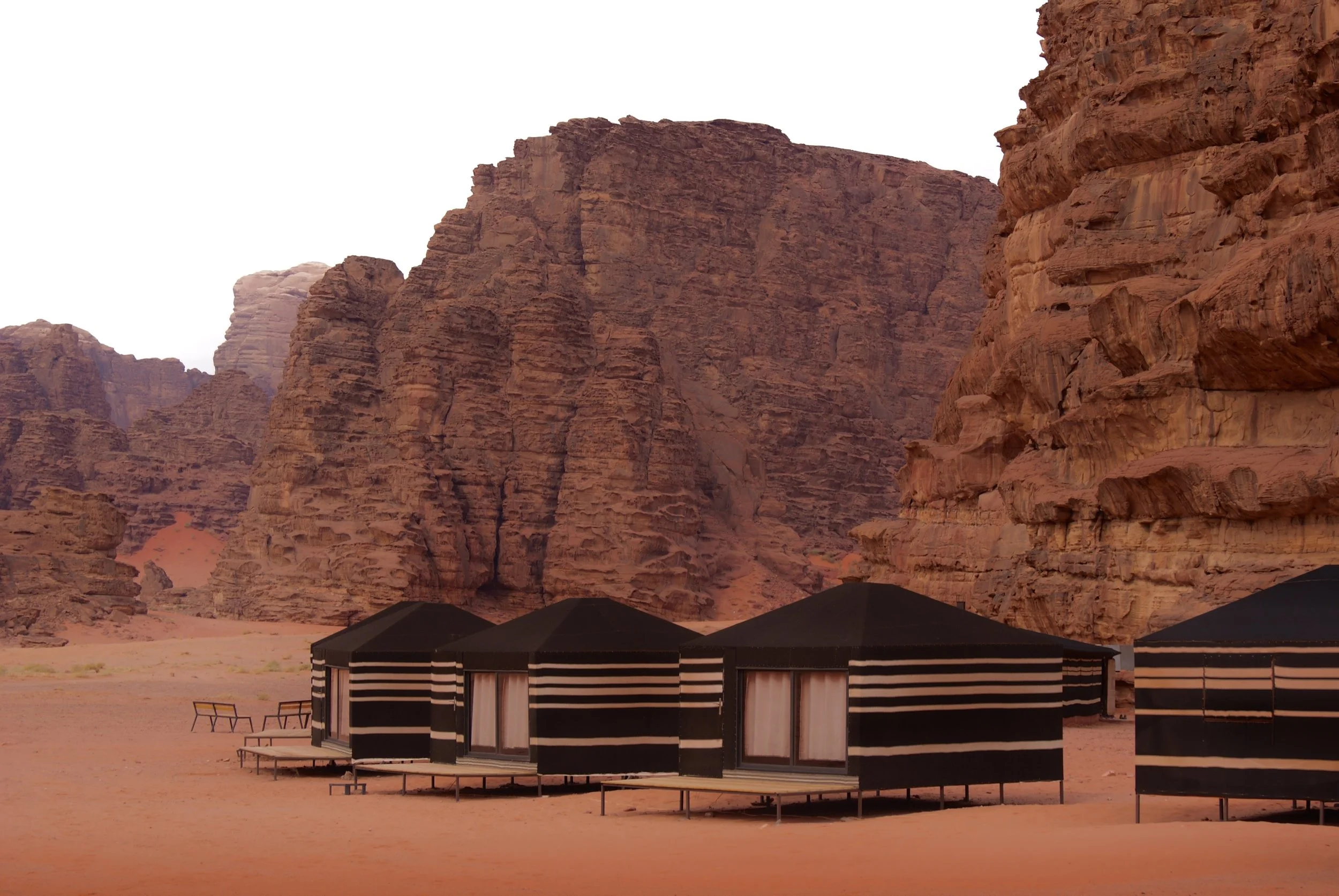 Four black and white striped tents set up on desert sand with massive red rock cliffs in the background. In Wadi Rum, Jordan.