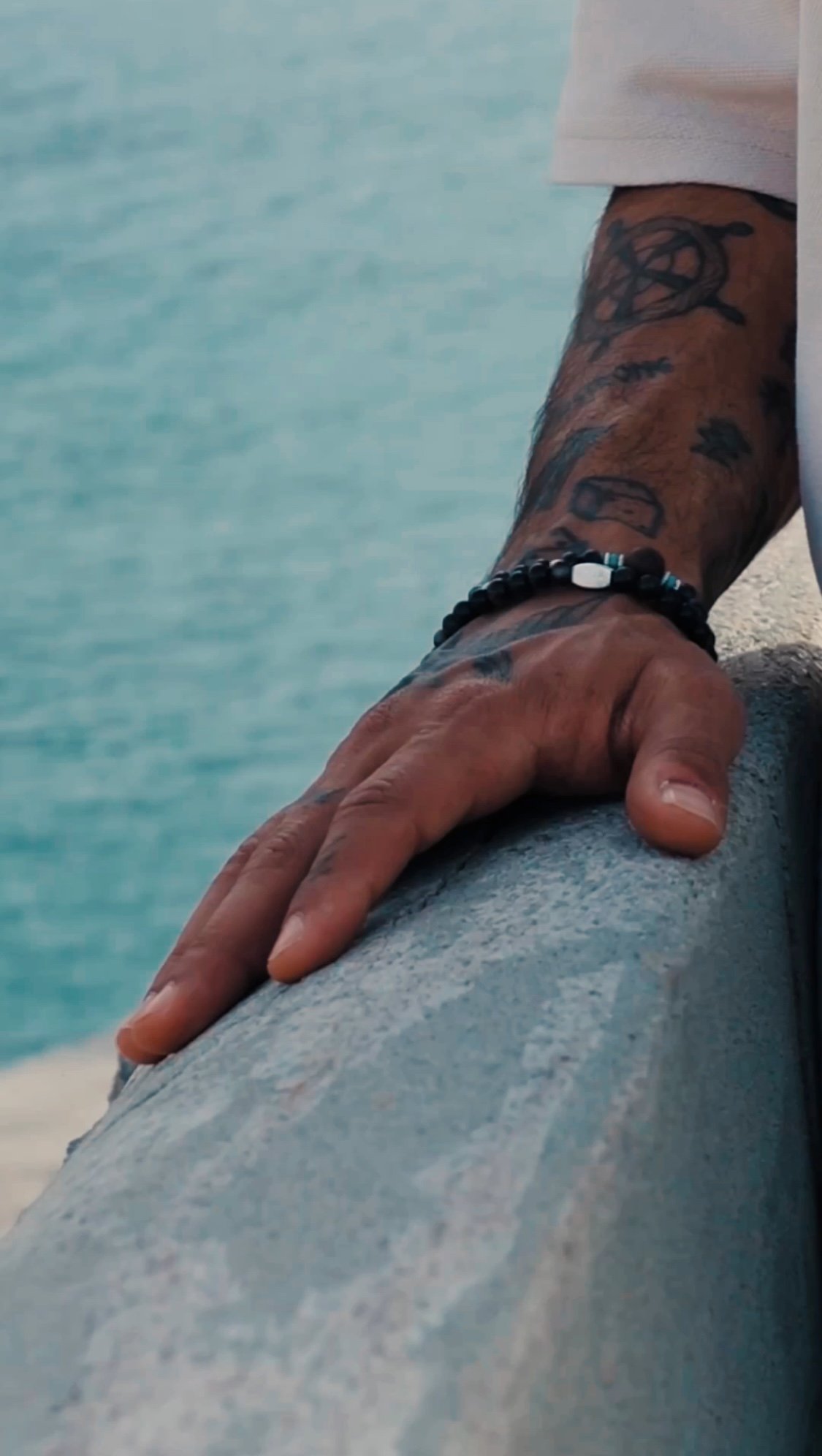 Close-up of a person's hand resting on a concrete surface, with a beard and tattoos on the arm, against a background of blue water.
