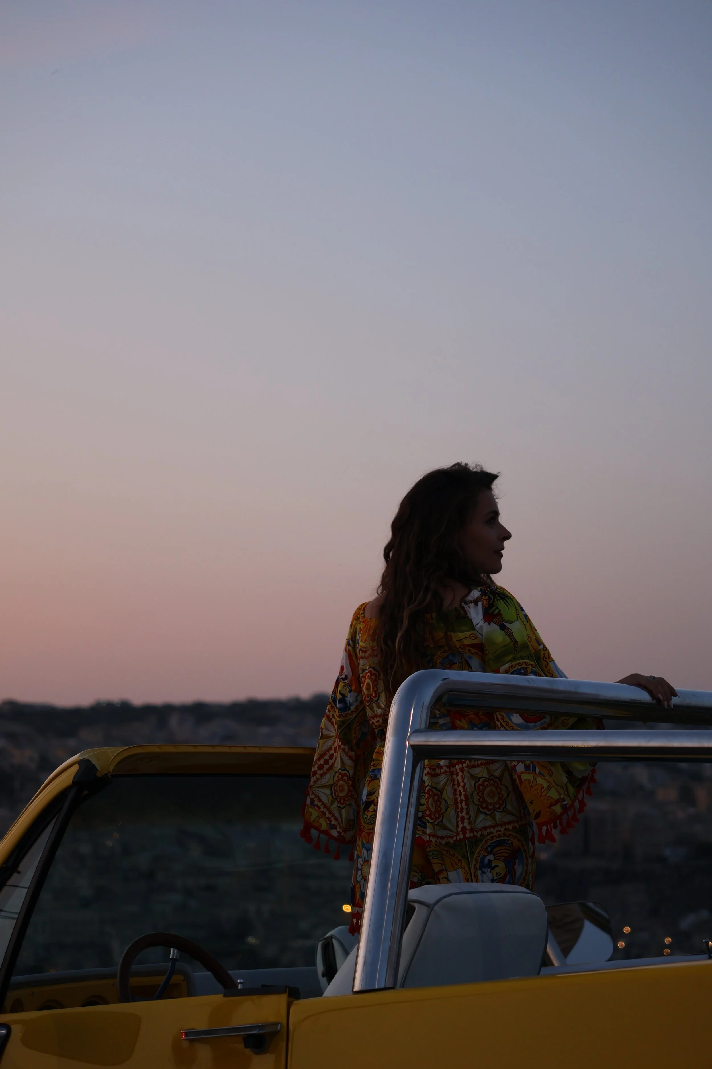 Person standing in a yellow convertible car at sunset or dusk, with a colorful patterned shirt, looking to the side, with a landscape background.