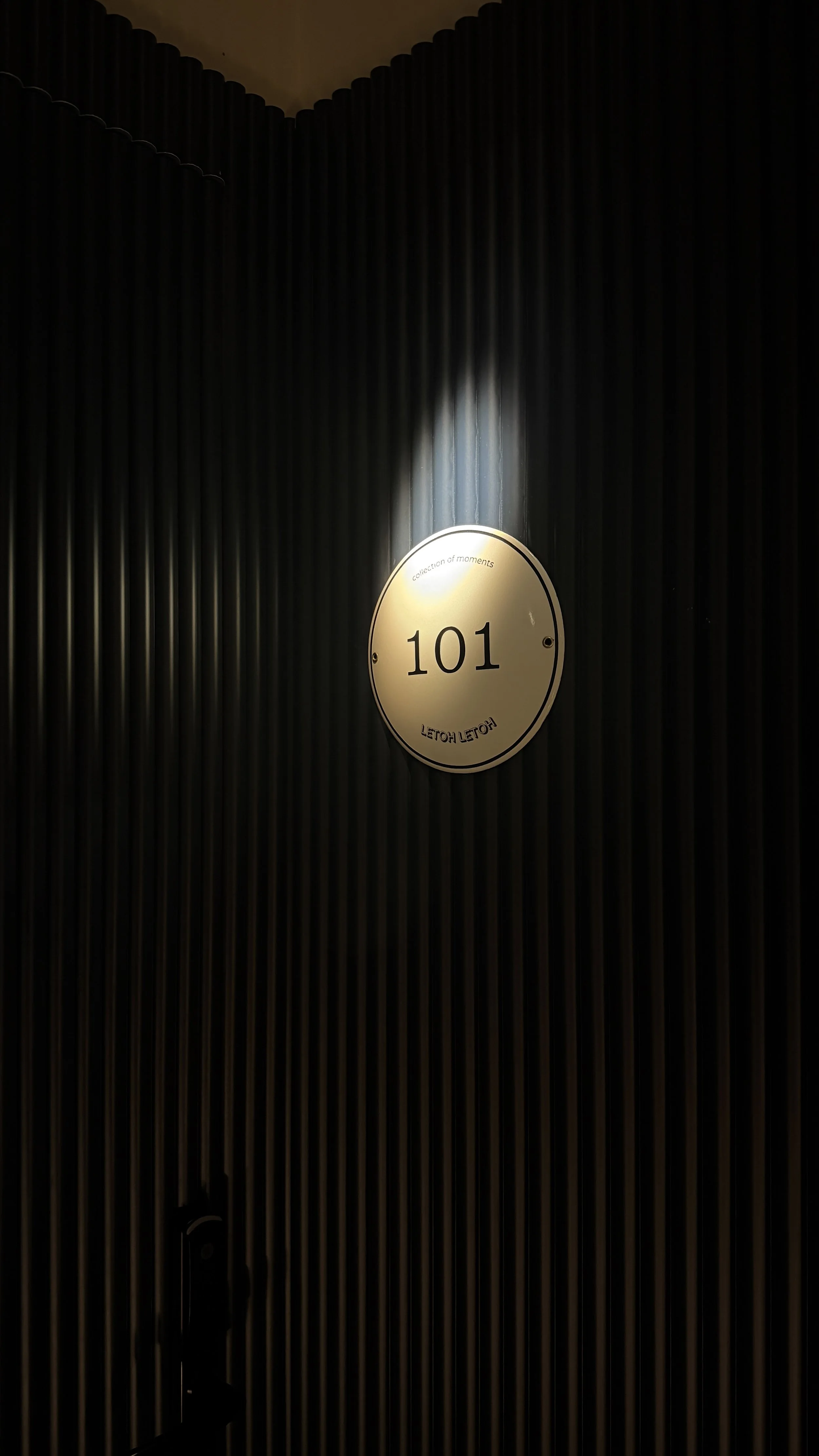 Close-up of a circular room number sign reading '101', mounted on a dark, ribbed wall with a spotlight shining on it.