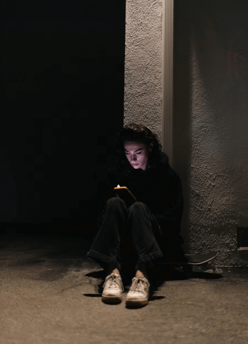 Persona sentada en el suelo contra una pared, iluminada por la luz del móvil en un entorno nocturno