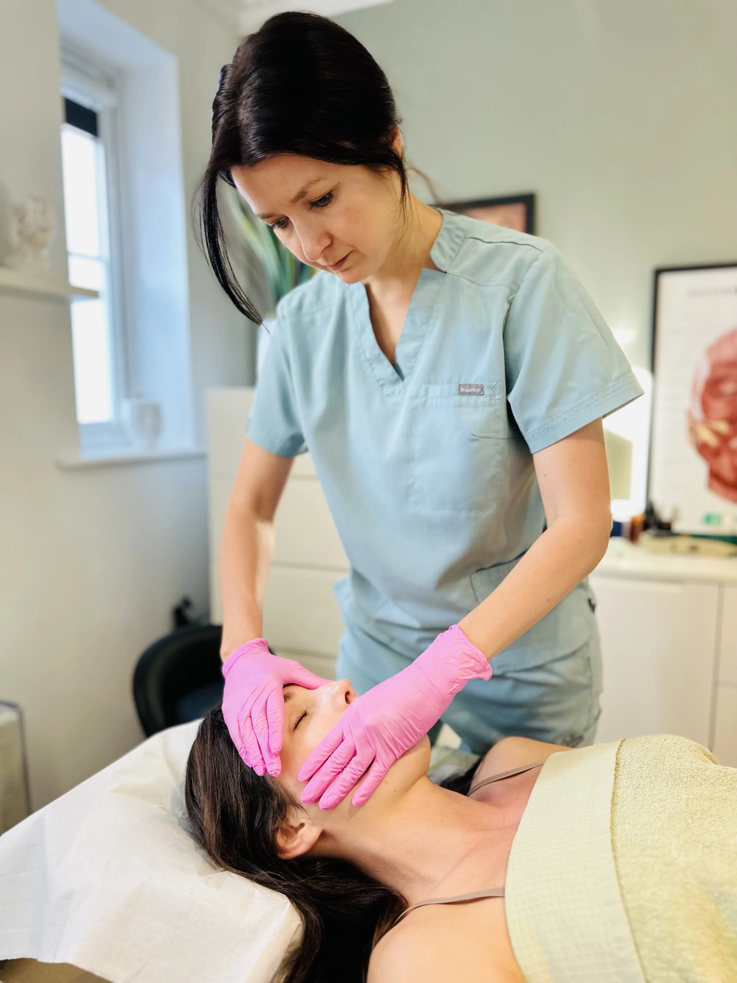 A healthcare professional wearing pink gloves performing a skincare treatment or examination on a woman lying on a treatment bed in a medical office.