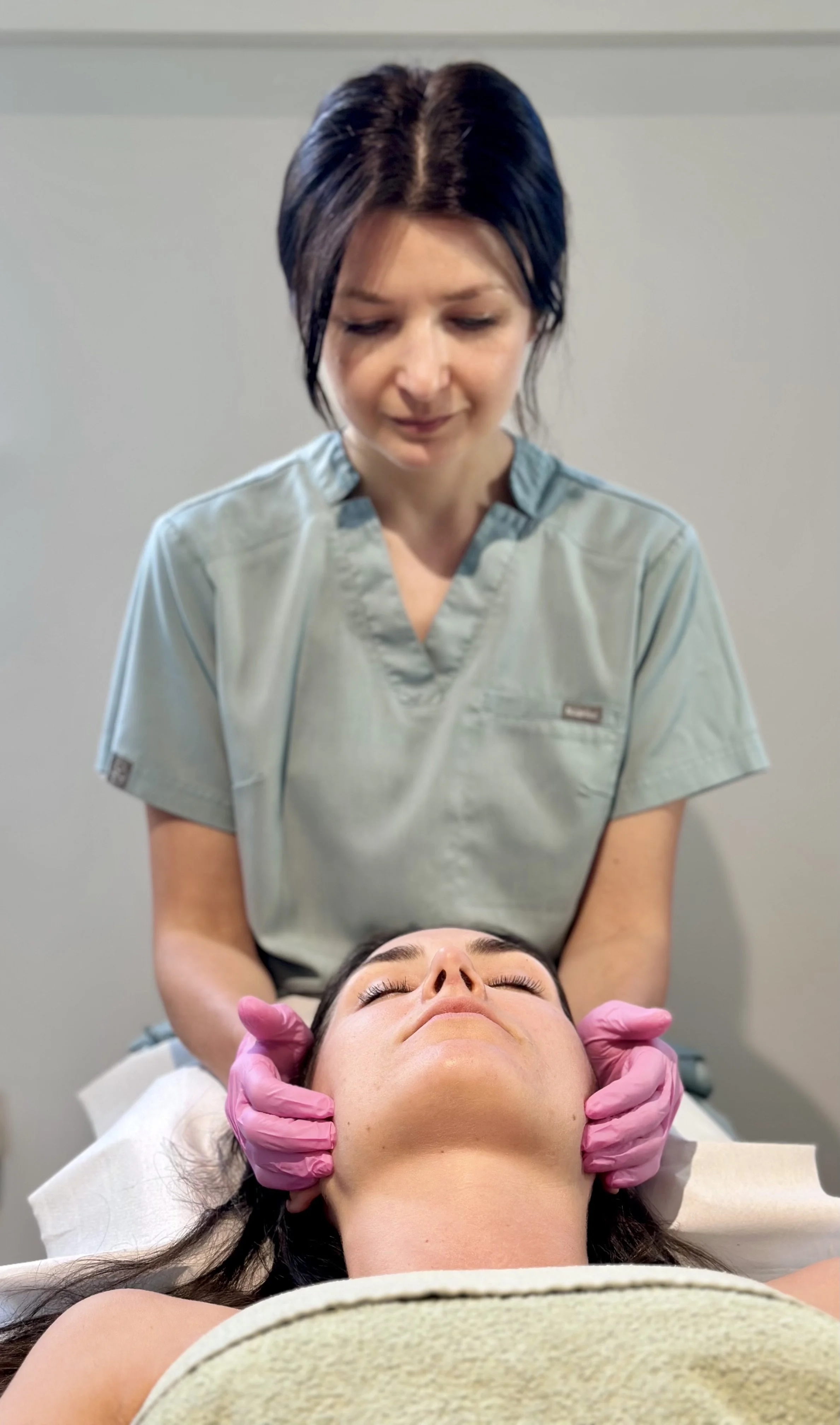 Medical professional in scrubs performs neck massage on patient lying down with eyes closed.