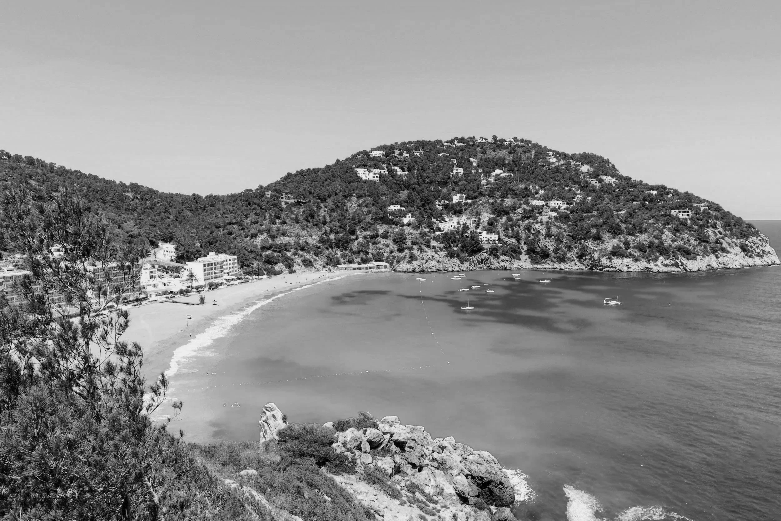 Strand met witte gebouwen en groene heuvels onder een blauwe hemel, met boten op het heldere blauwe water.