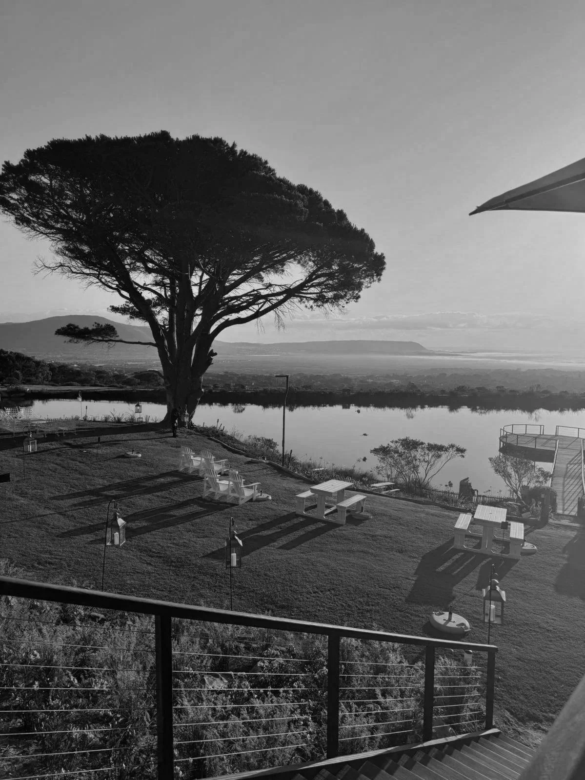 Uitzicht over een tuin met zitplaatsen, een grote boom en een meer, bij zonsondergang.