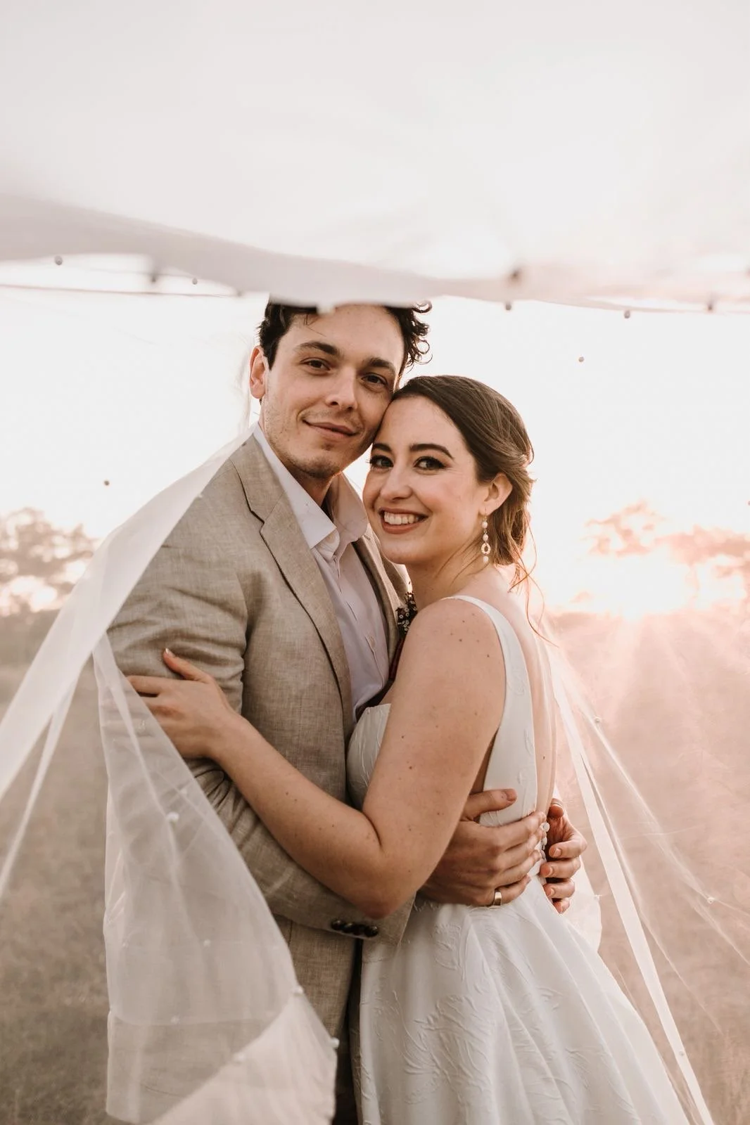 A newlywed couple standing outdoors at sunset, smiling and hugging, with the bride wearing a white wedding dress and the groom in a beige suit, under a sheer fabric canopy.