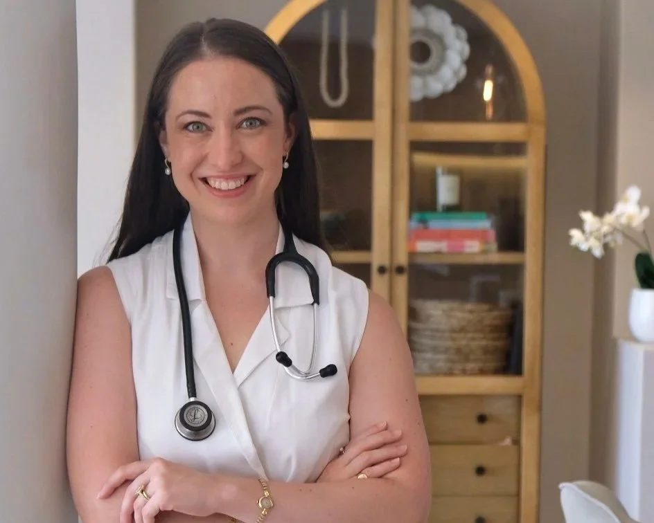 A smiling woman with dark brown hair, wearing a white sleeveless top and a stethoscope around her neck, stands with arms crossed in a room with wooden furniture and a flower in a vase.