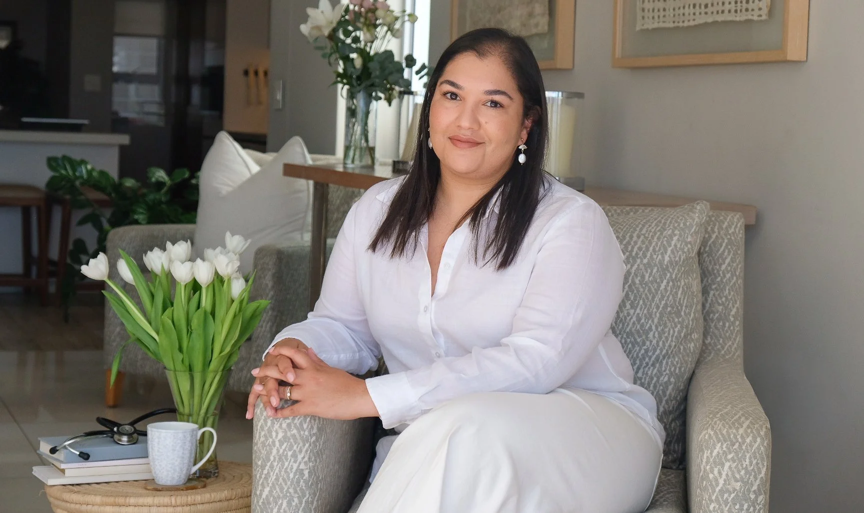 A woman with dark hair dressed in white, sitting on a beige patterned chair in a living room with a bouquet of white tulips on a small table beside her.