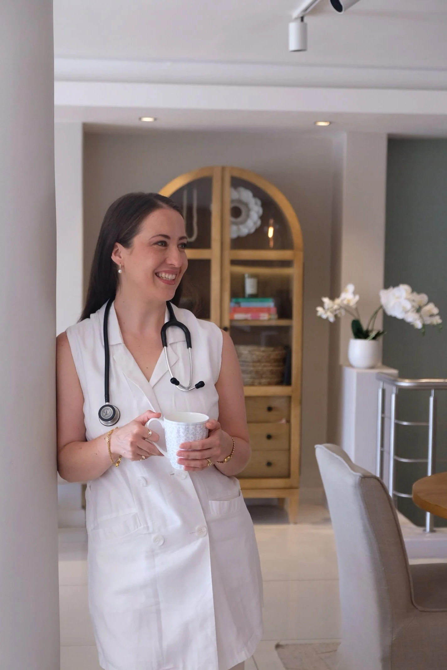 A South African doctor wearing a white sleeveless coat and stethoscope, smiling and holding a mug in a cozy, well-lit room with decor including a wooden shelf, flowers, and dining chairs.