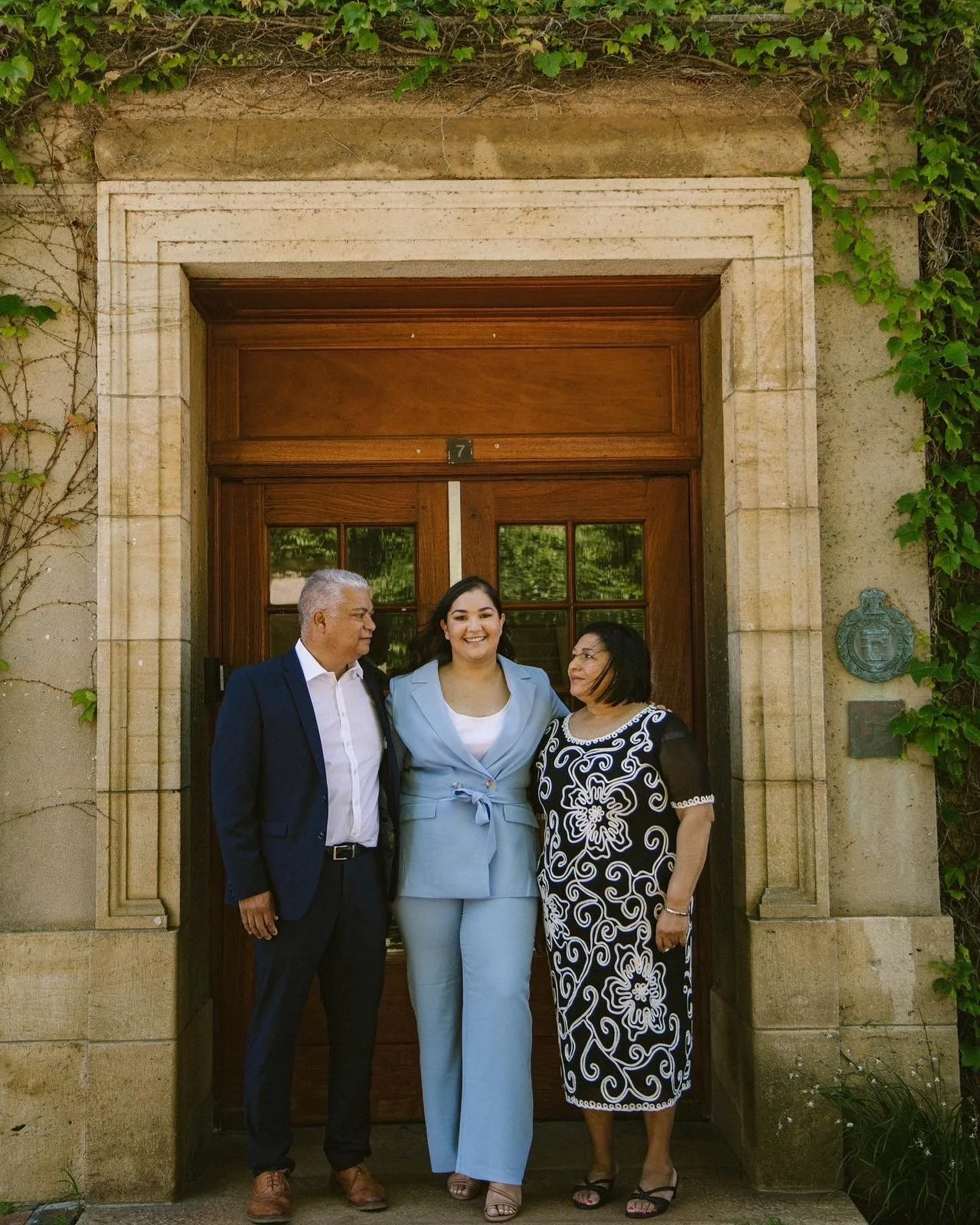 Three people standing in front of a wooden door, posed for a photo, with a stone frame and greenery around.