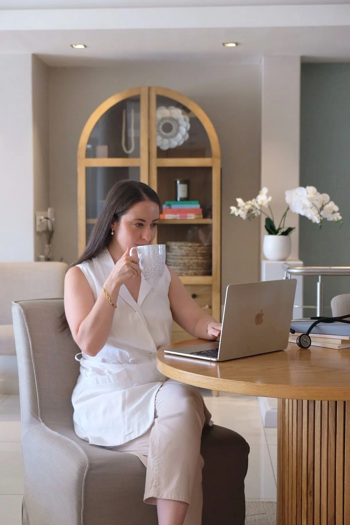 A South African woman doctor in white sleeveless outfit sitting at a dining table, drinking from a mug while working on a silver laptop in a cozy, modern home with wooden and white decor.