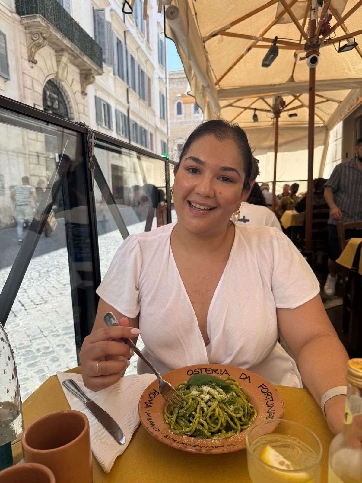 A smiling woman dining outdoors at a restaurant with a plate of green pasta in front of her. She is holding a fork and wearing a light-colored top. The restaurant has yellow tablecloths, umbrellas, and a glass window showing a cobblestone street and historic buildings.