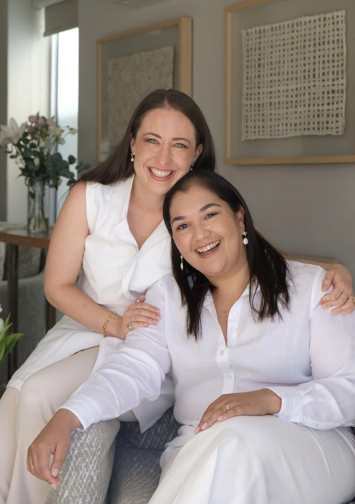 Two South African women doctors smiling and hugging in a living room, dressed in white, with artwork on the wall and a vase of flowers in the background.