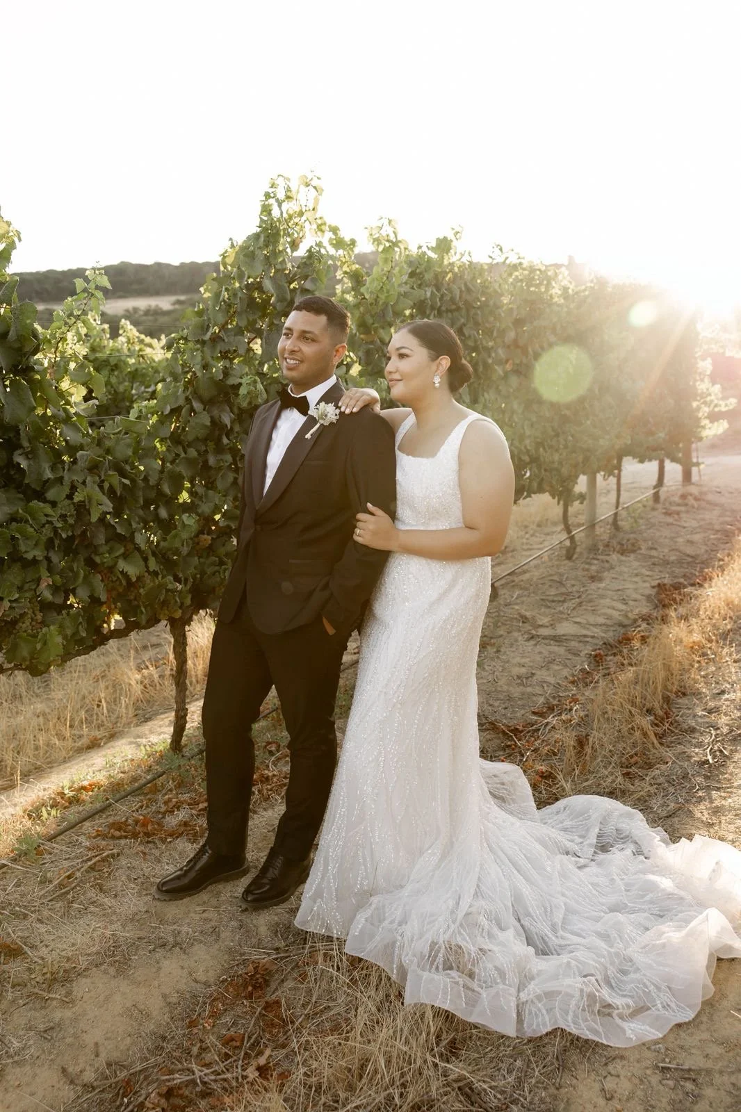 A newlywed couple in wedding attire posing in a vineyard during sunset, with the bride in a white gown and the groom in a black tuxedo.