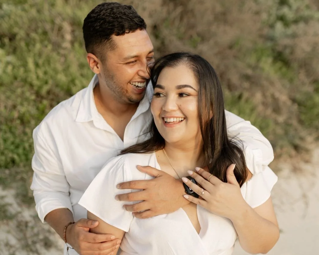 A smiling couple in white clothing enjoying a moment outdoors, with a man embracing a woman from behind.