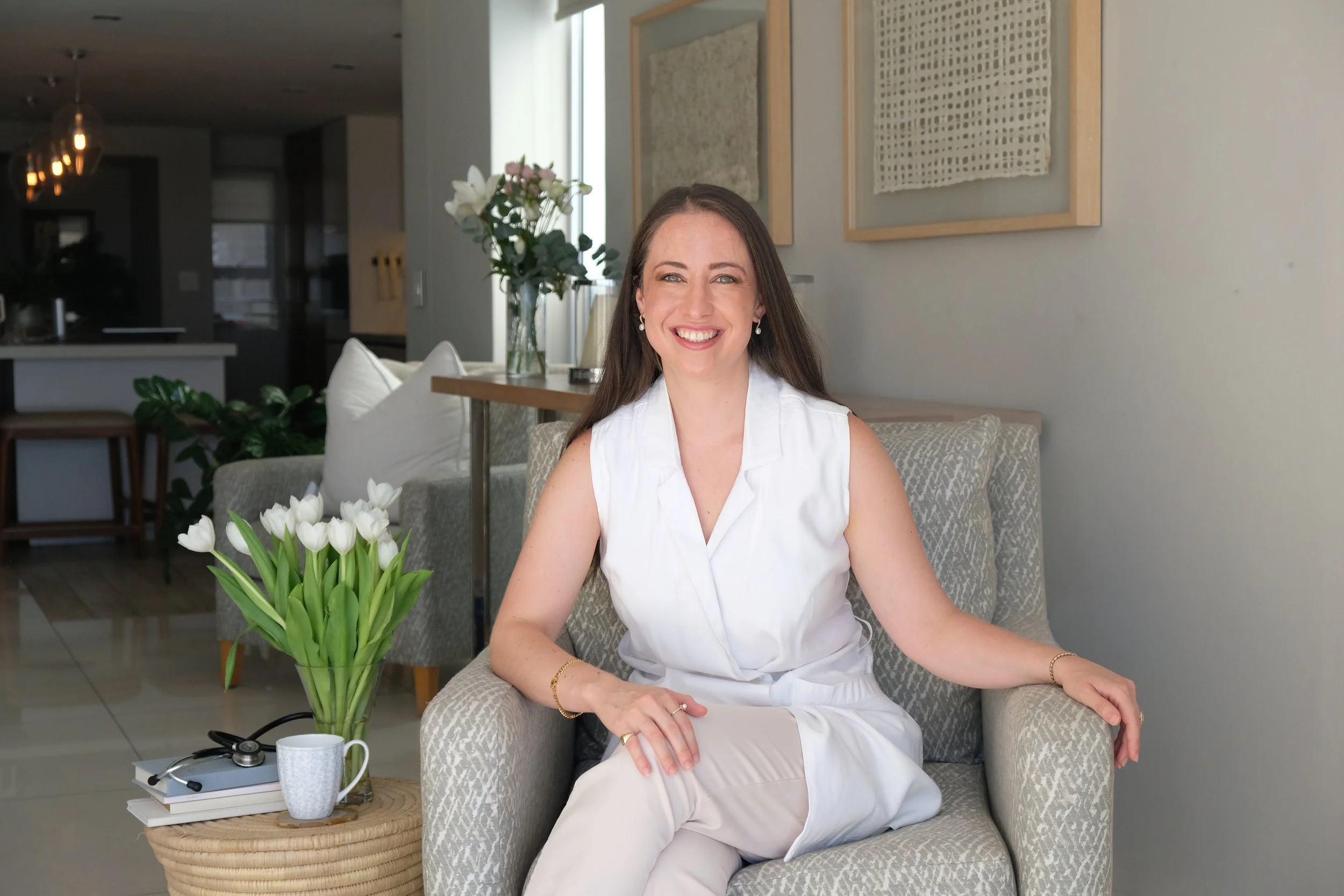 A woman with long brown hair smiling and sitting in an armchair in a modern living room with white walls, contemporary artwork, a vase of white tulips on a small round table, and a kitchen in the background.