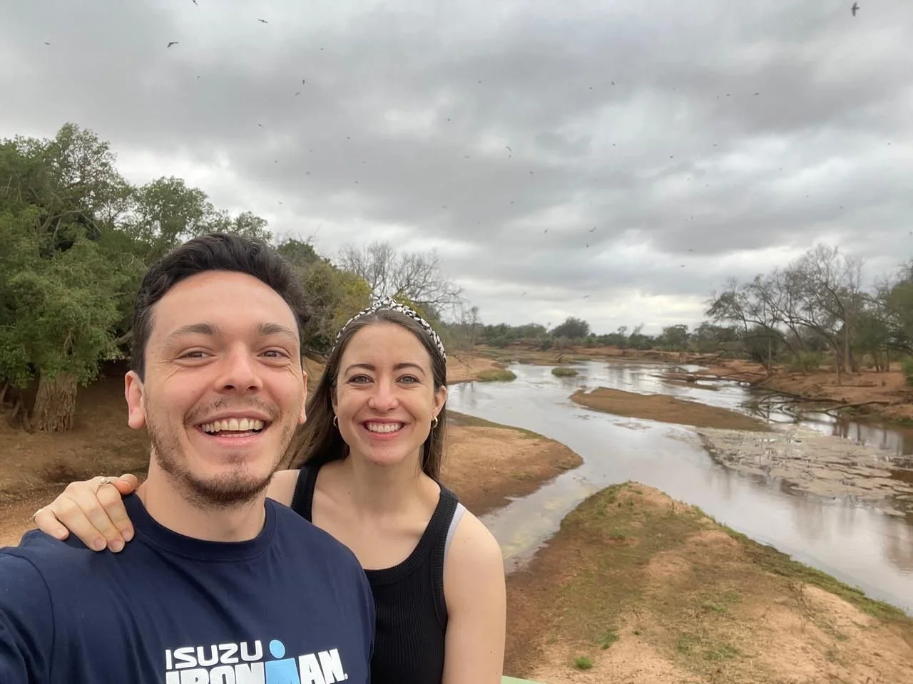 A smiling man and woman taking a selfie outdoors near a river on a cloudy day.
