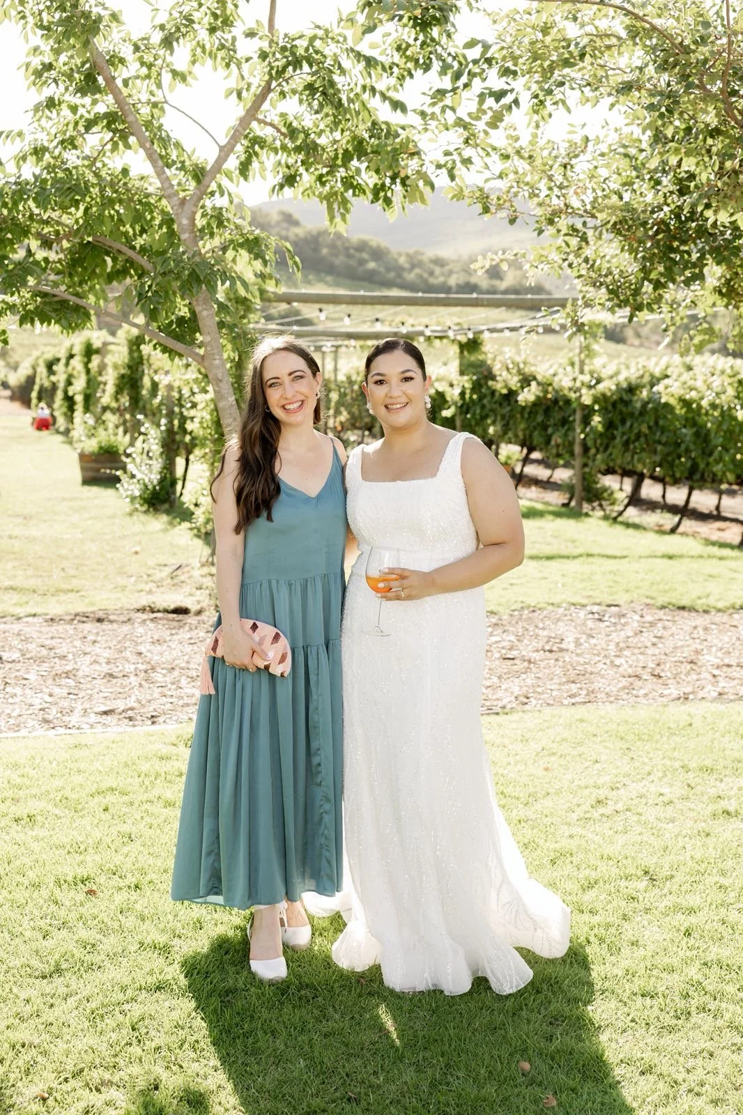 Two women standing outdoors in a vineyard, smiling at the camera. One woman wears a blue dress and holds a clutch, the other wears a white dress and holds a glass of wine.