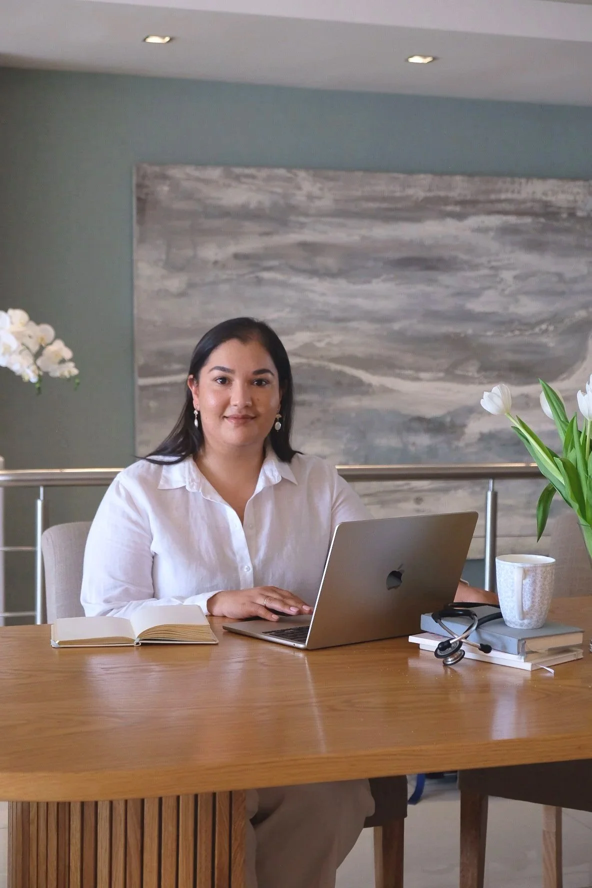 A woman with dark hair and earrings, wearing a white shirt, sits at a wooden table with a silver laptop, an open notebook, a stack of books with a stethoscope on top, a cup of coffee, and a vase of white tulips. She is in a modern, well-lit room with an abstract painting behind her.