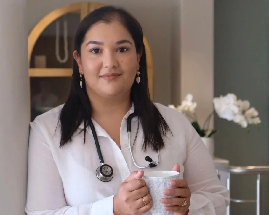 A woman with dark hair, wearing a white shirt and earrings, holding a mug in a room with a bookshelf and white flowers in the background.