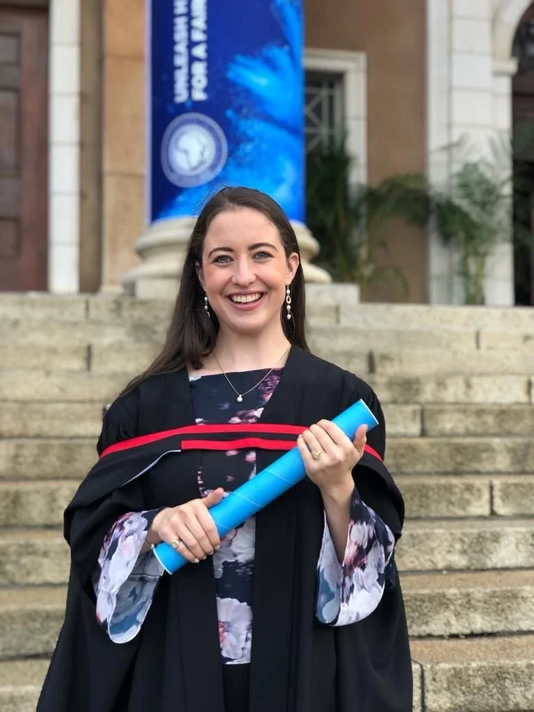 A young woman in graduation cap and gown holding a diploma, standing on stone steps outside a building with a banner in the background.