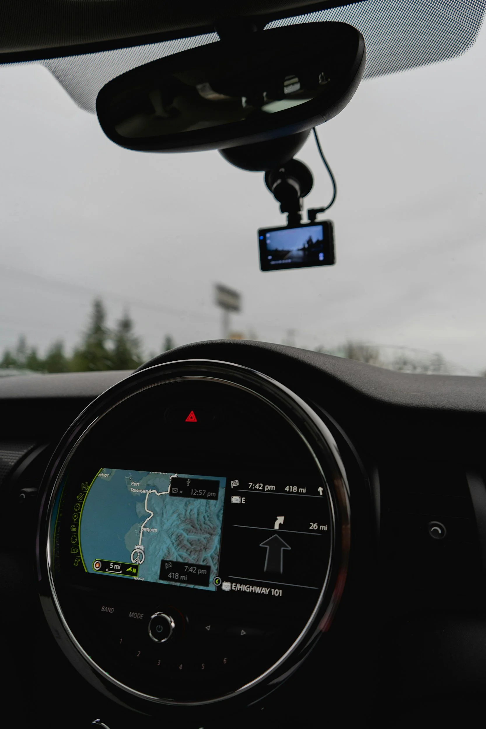 Interior de un coche mostrando el tablero de navegación y la cámara en el espejo retrovisor. La pantalla del GPS indica que el coche está en la autopista 101, a 418 millas del destino, con la dirección ascendente. La cámara del dash cam está instalada en el espejo retrovisor, grabando la vista del exterior en un día nublado.