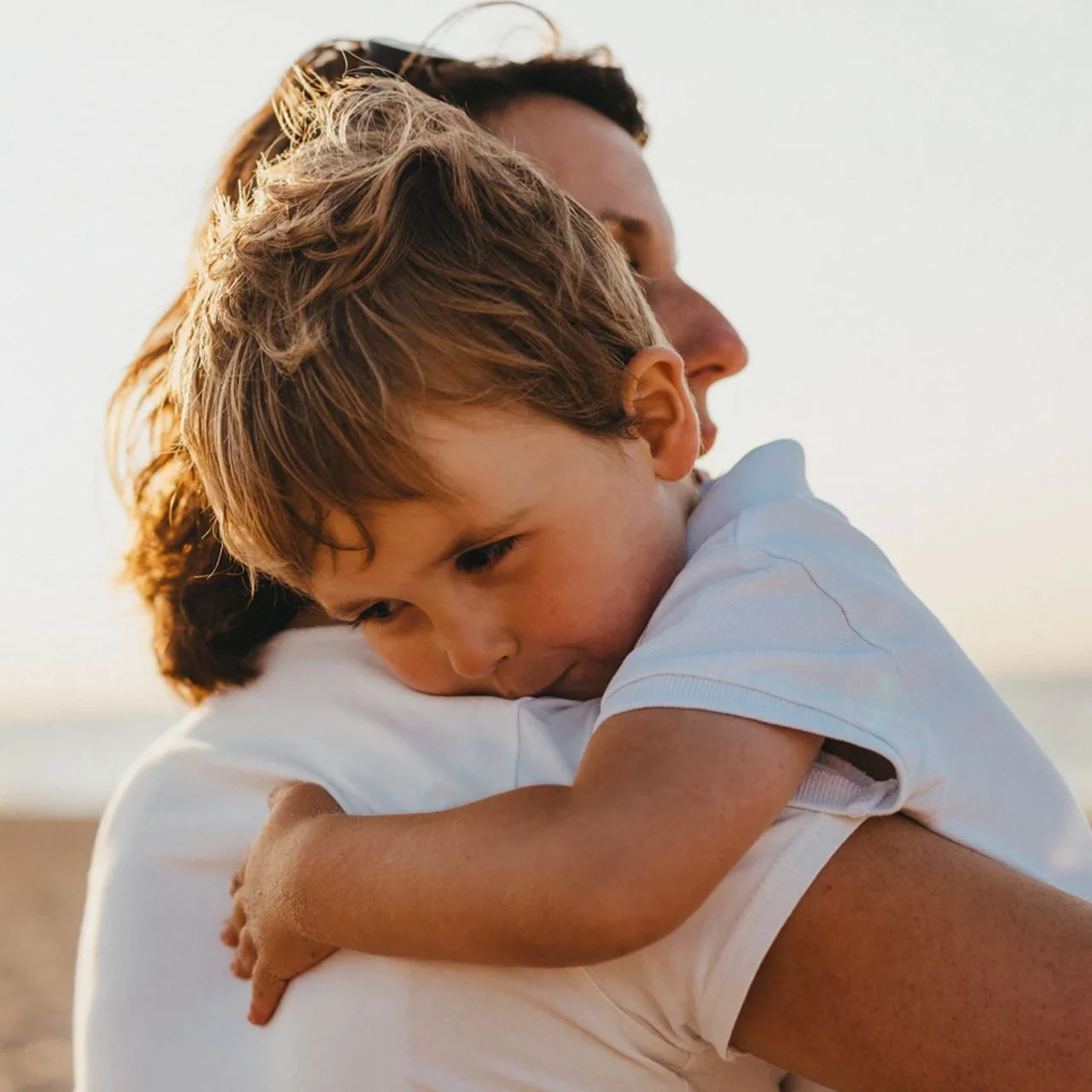 Un niño pequeño abrazando con nostalgia a un adulto en una playa al atardecer.