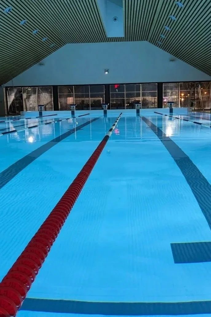 Piscine intérieure vide avec des couloirs de nage, éclairage tamisé et fenêtres donnant sur l'extérieur.