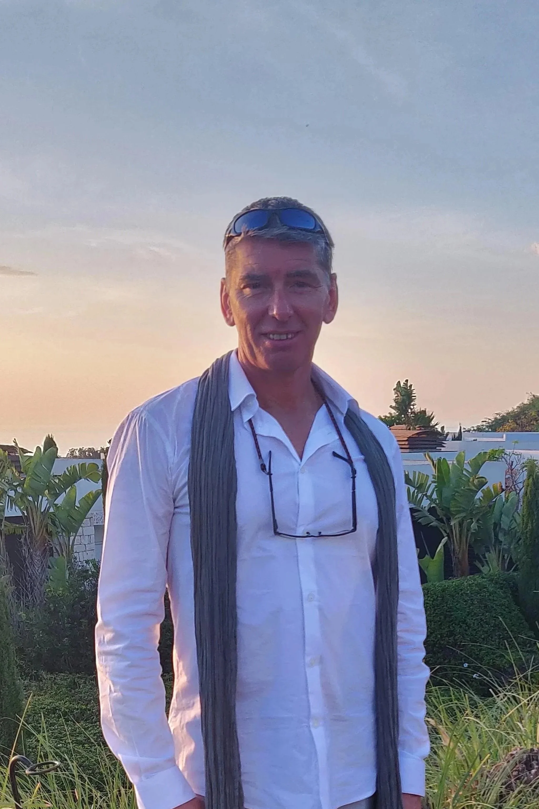 Un homme portant une chemise blanche, avec des lunettes autour du cou, des lunettes de soleil sur la tête, se tient dans un jardin avec des plantes tropicales et un ciel au coucher du soleil.