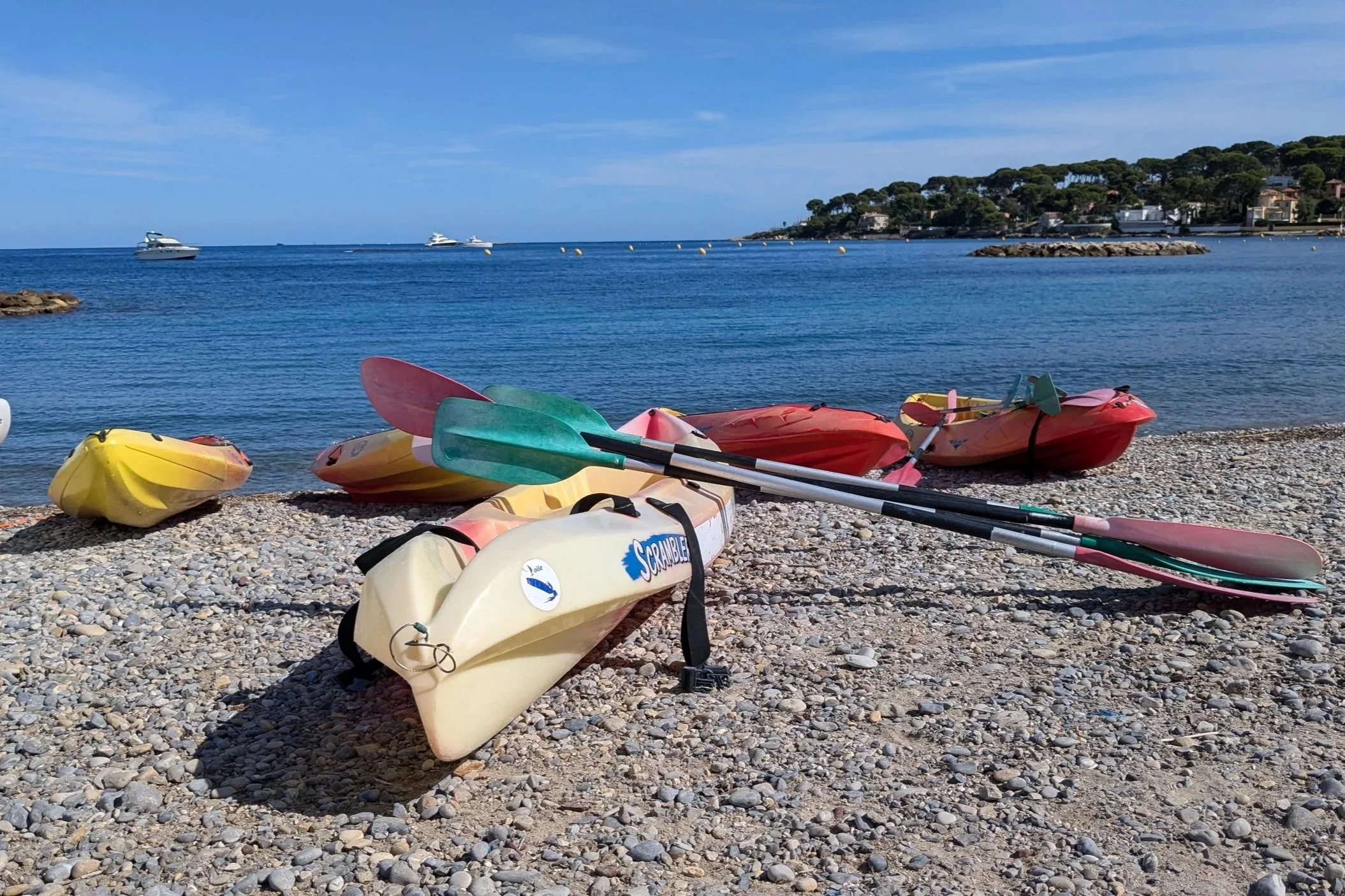Kayaks et paddles sur une plage de galets au bord de la mer, avec yachts et villas en arrière-plan sous un ciel ensoleillé.