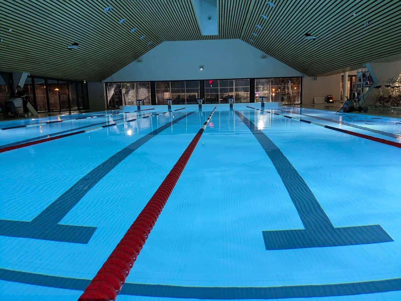 Piscine intérieure vide avec de la lumière bleue, lignes de nageur, murs vitrés et équipement de fitness dans le fond.